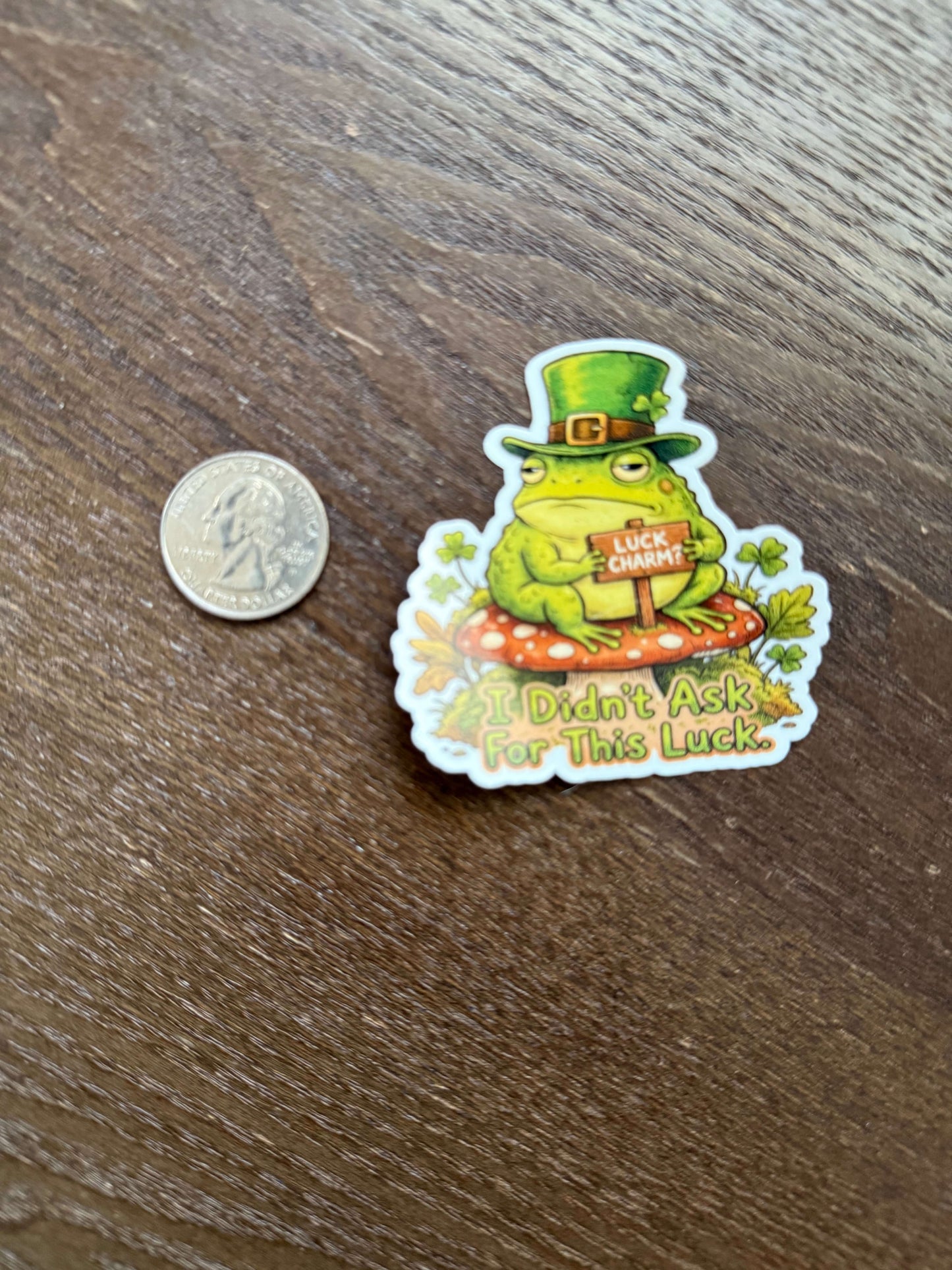 Sticker of a frog wearing a top hat with text on a wooden surface next to a coin for size reference.