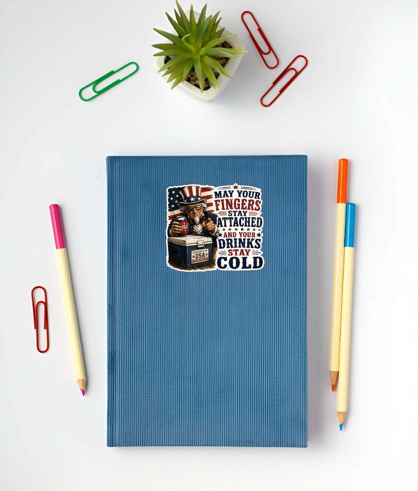 Blue notebook with a sticker featuring a detailed vintage woodcut illustration of Uncle Sam wearing flag sunglasses, holding a USA beverage can next to a blue ice chest labeled "PROPERTY OF USA", alongside bold red and blue text reading "MAY YOUR FINGERS STAY ATTACHED AND YOUR DRINKS STAY COLD". on a white surface with stationery items.