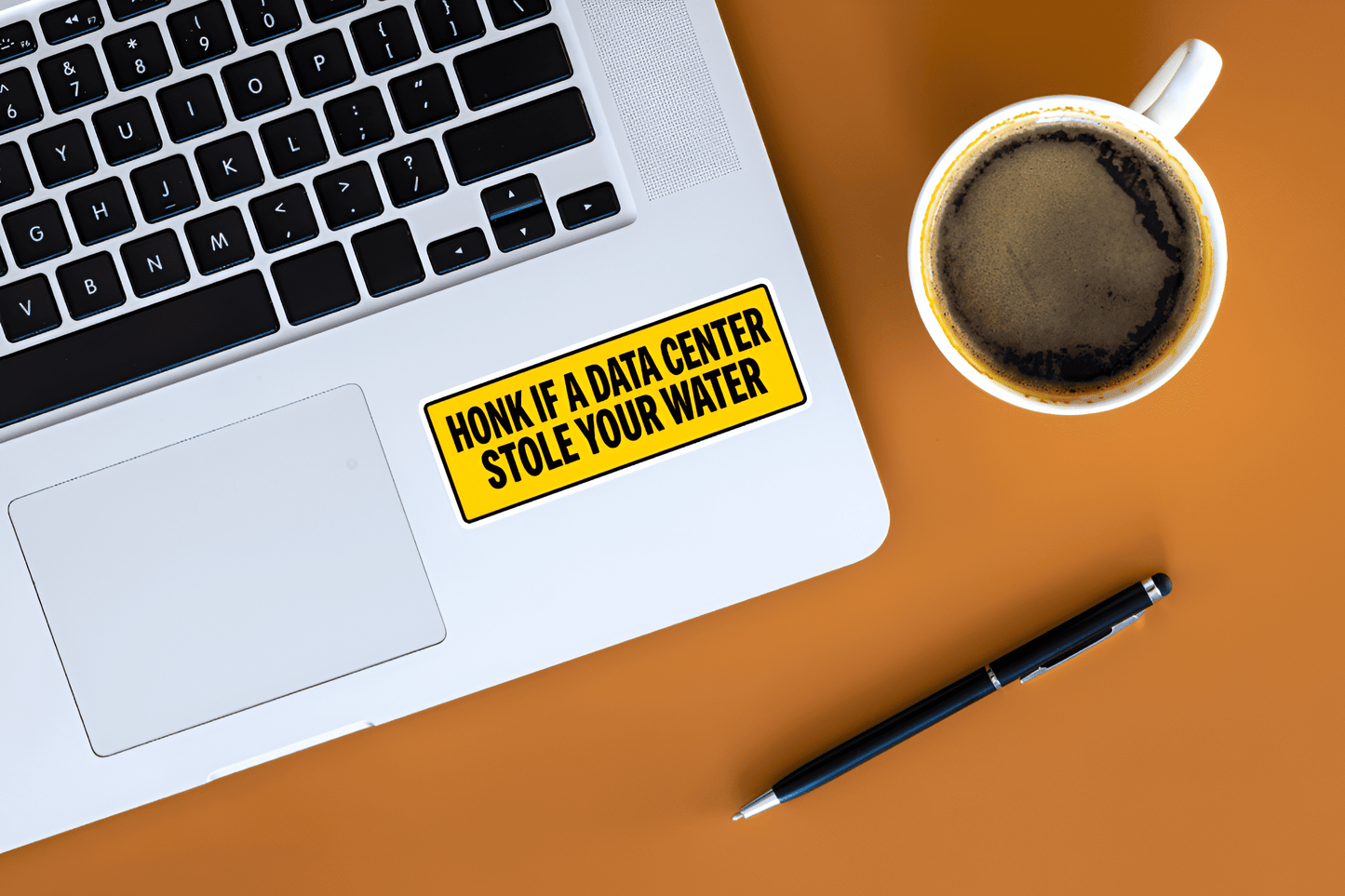 Laptop with a humorous sticker on a desk with a coffee cup and pen.