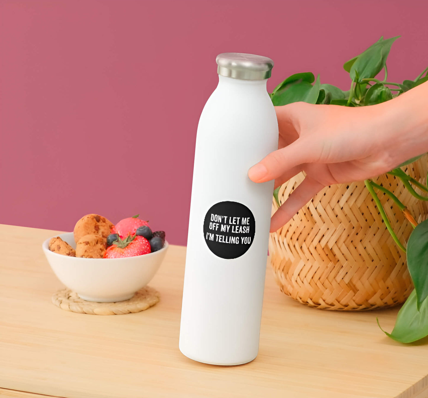 White water bottle with a black label held by a hand on a wooden table with a bowl of snacks and a plant in the background.