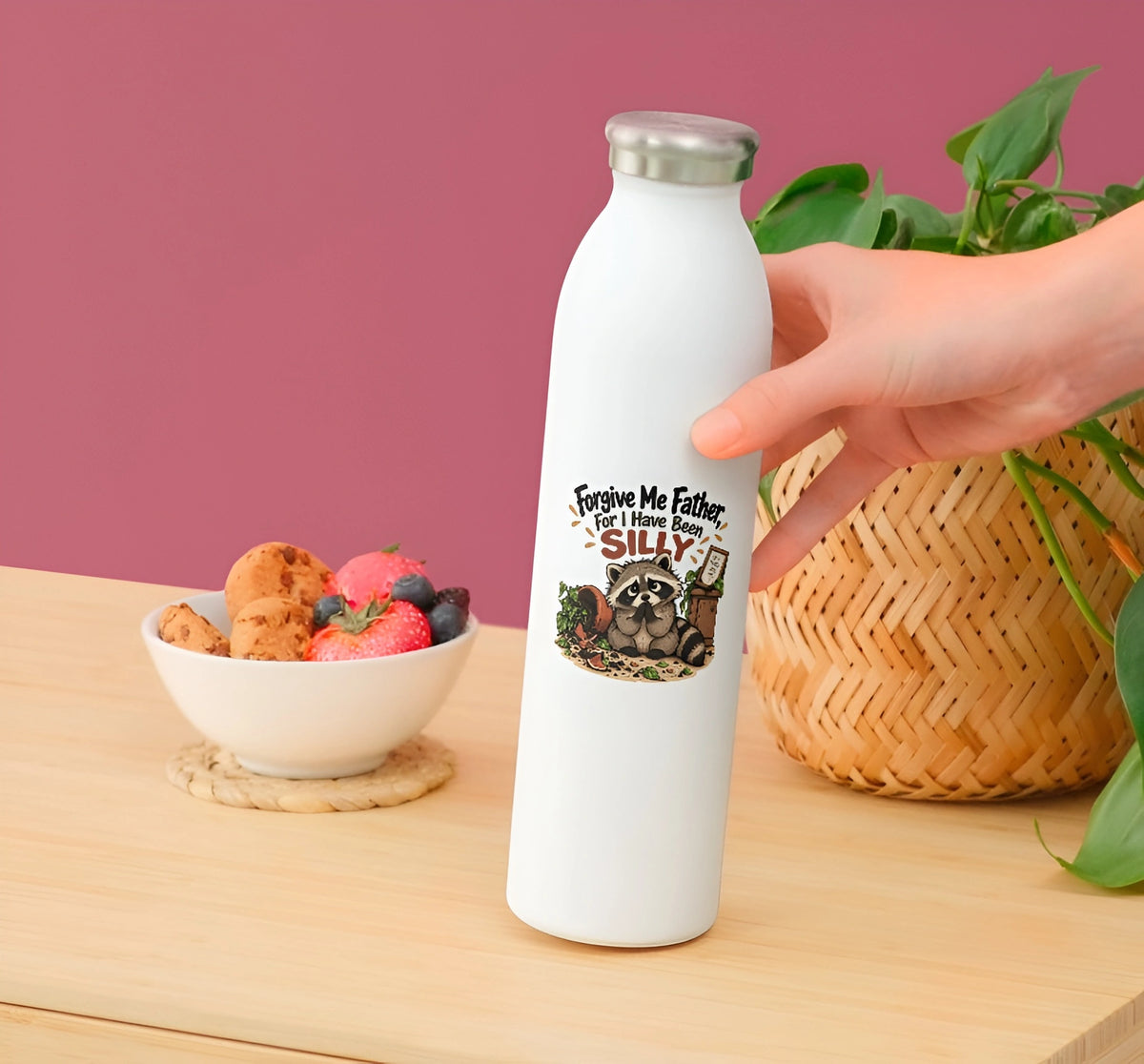 A hand holding a white insulated water bottle with a silver cap, featuring a raccoon graphic and the text 'Forgive My Father, For I have been Silly' on a wooden table. A bowl of fruit, a woven basket, and green leaves are in the background against a dusty pink wall.