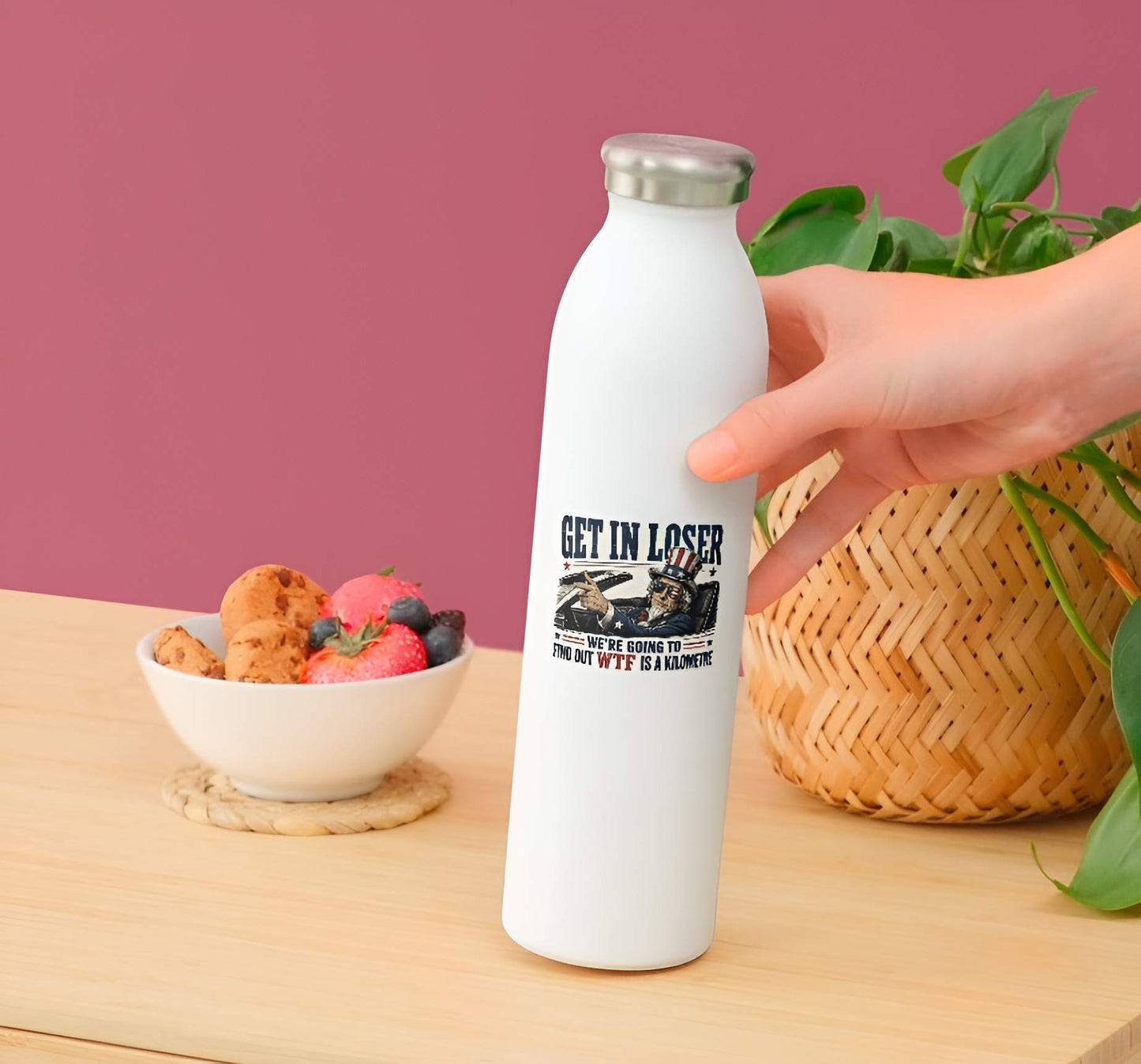 White water bottle with a sticker featuring a detailed vintage-style illustration of Uncle Sam wearing flag sunglasses and driving a convertible car, pointing forward next to bold text reading "GET IN LOSER WE'RE GOING TO FIND OUT WTF IS A KILOMETRE". on a wooden table with a bowl of fruit and plant in the background.