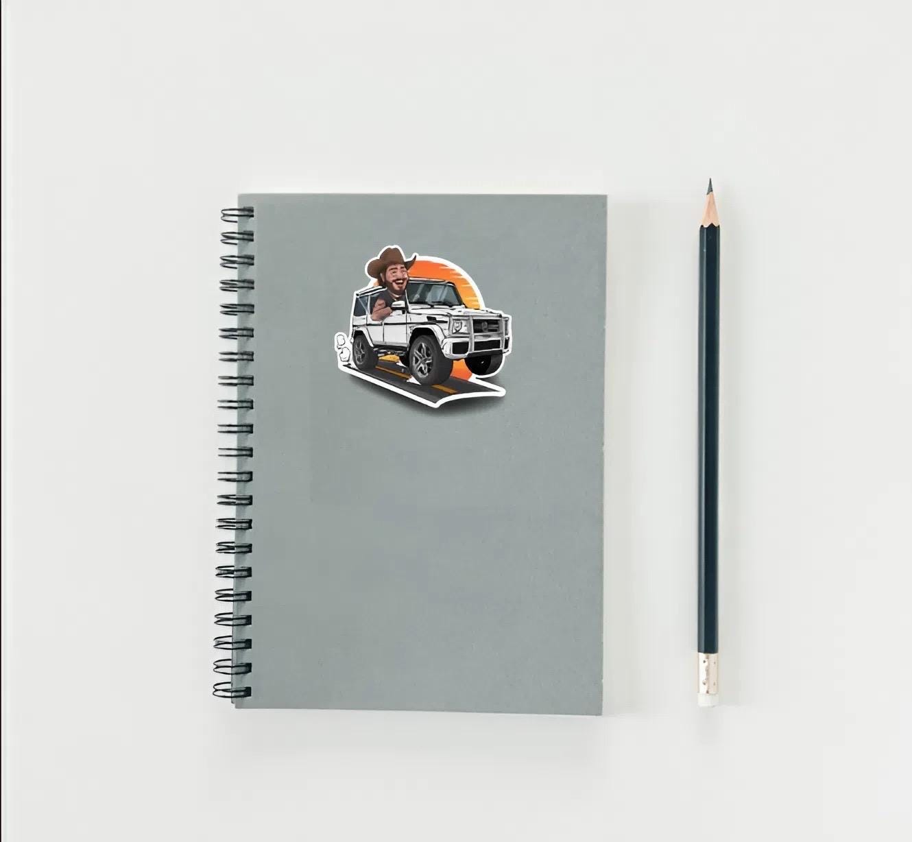 Notebook with a sticker of a Post Malone in a cowboy hat driving a car, next to a pencil on a light gray background