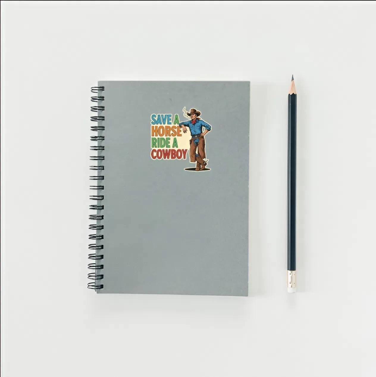Gray notebook with cowboy illustration and text, accompanied by a pencil on a light gray background