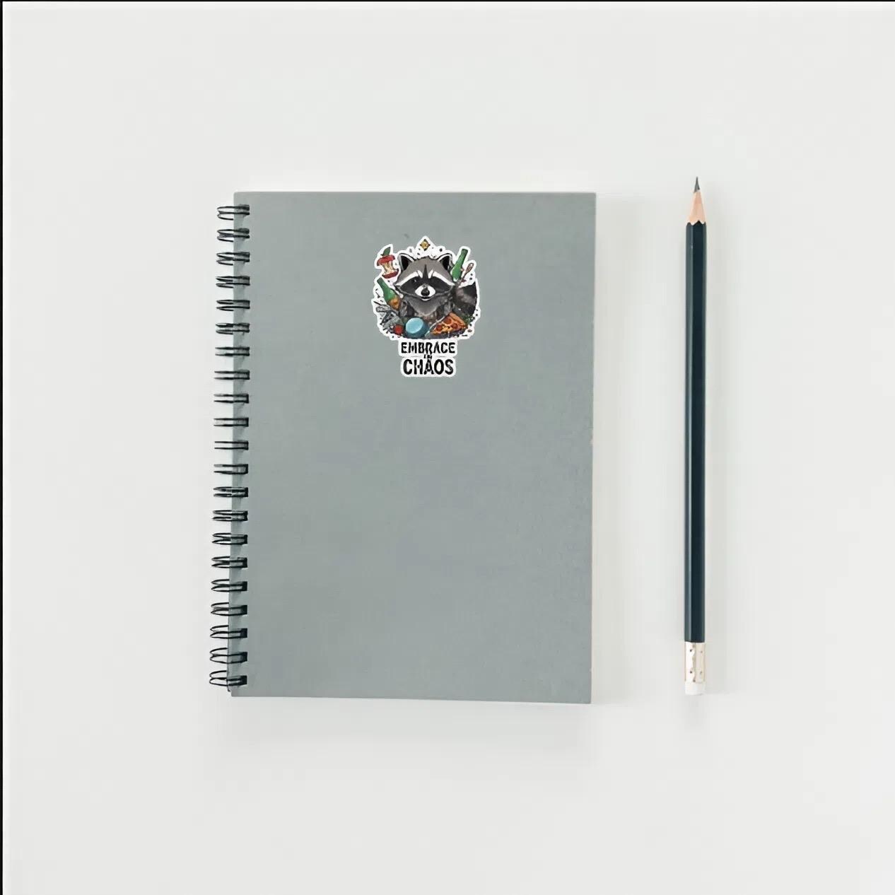 Gray notebook with a raccoon illustration and text, accompanied by a black pencil on a white background.