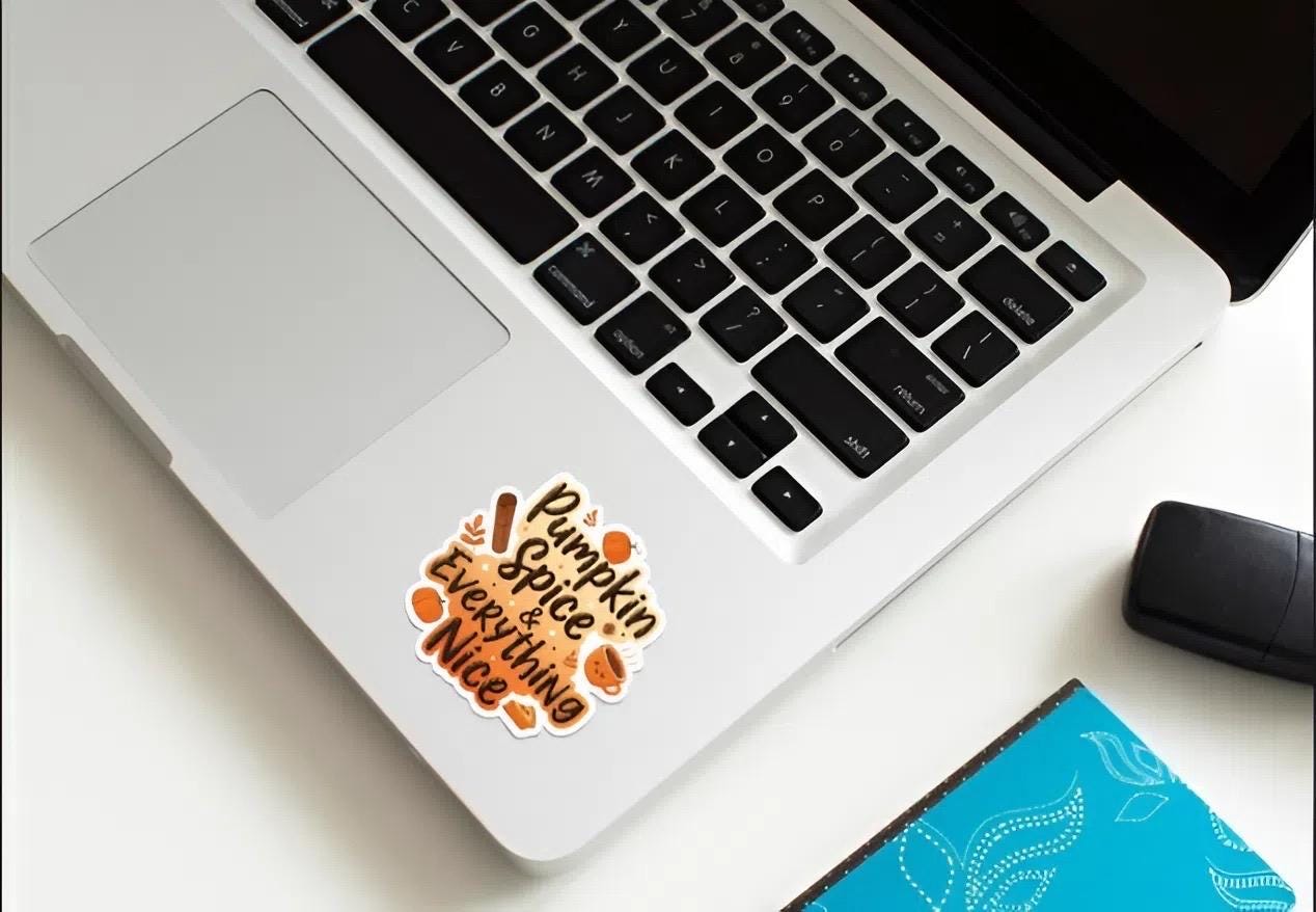 Laptop with a 'Pumpkin Spice & Everything Nice' sticker on a white surface with a mouse and notebook.