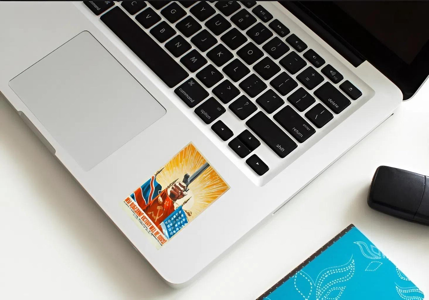 Laptop with a sticker on the palm rest area, next to a mouse and notebook on a light surface.