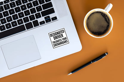 Laptop with a 'Designated Driver' sticker, coffee cup, and pen on an orange surface