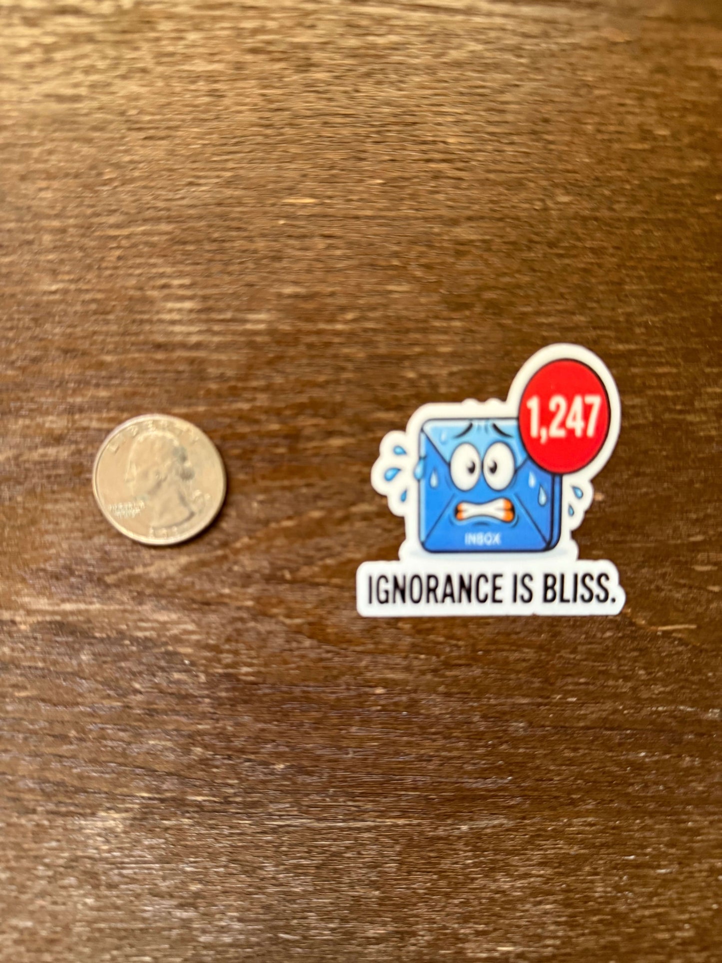 Sticker with a cartoon character and text 'IGNORANCE IS BLISS' next to a coin on a wooden surface