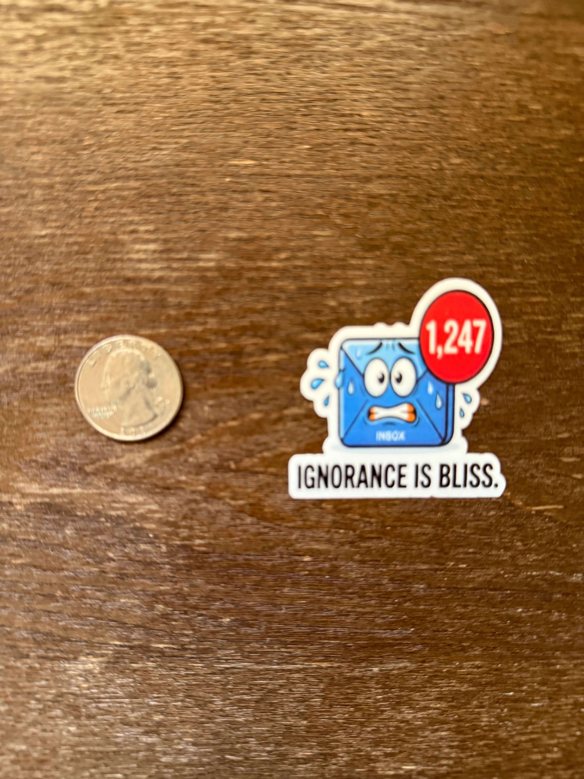 Sticker with a cartoon character and text 'IGNORANCE IS BLISS' next to a coin on a wooden surface