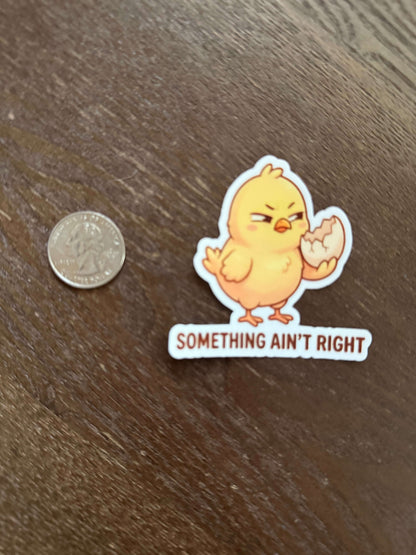 Sticker with a cartoon chick and text 'SOMETHING AIN'T RIGHT' on a wooden surface with a coin for scale.