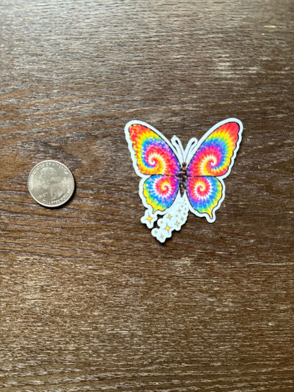 Colorful butterfly-shaped tie-dye sticker on a wooden surface with a coin for scale.