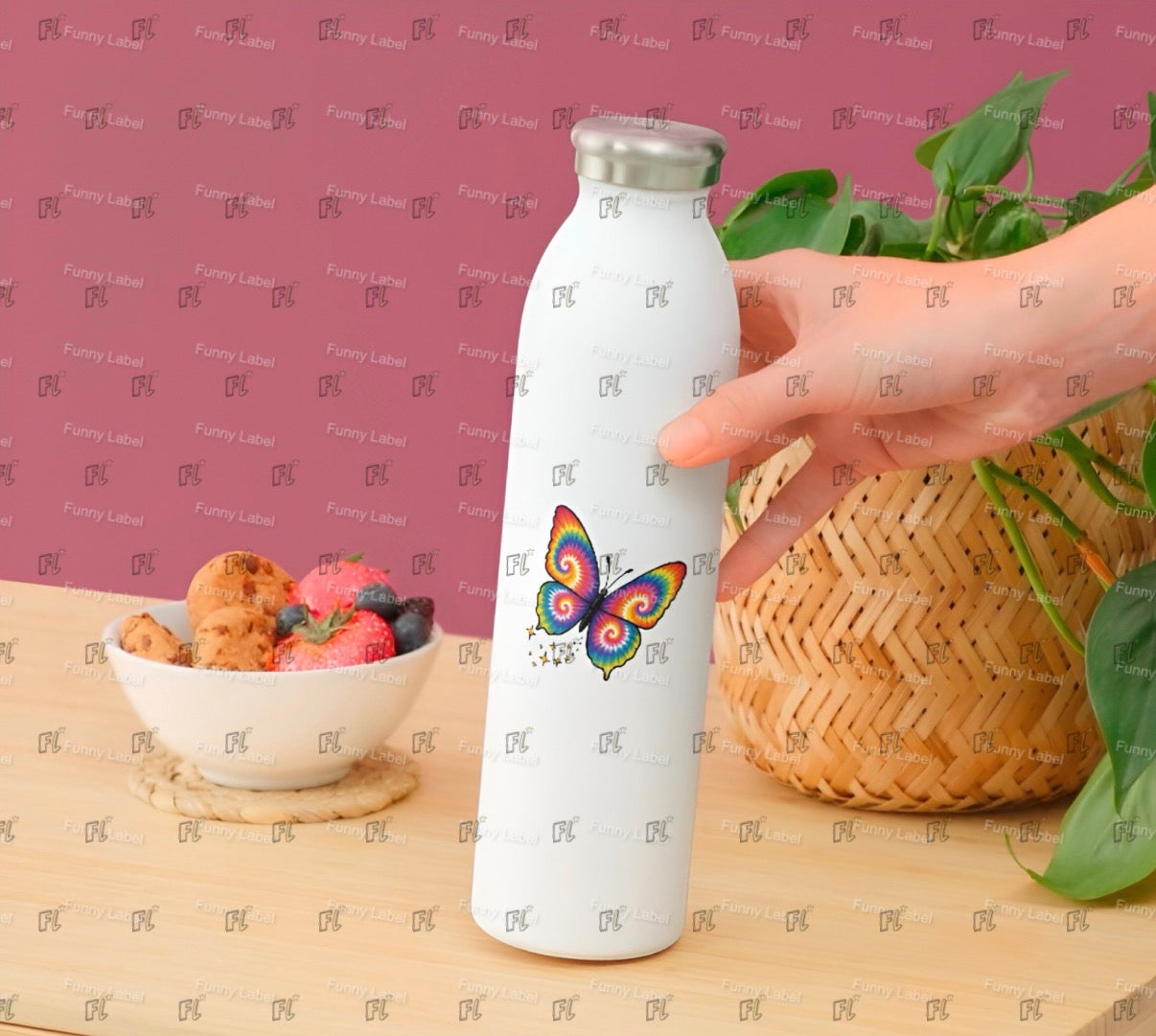 White water bottle with a colorful butterfly design held by a hand on a wooden surface with a bowl of fruit and basket in the background.