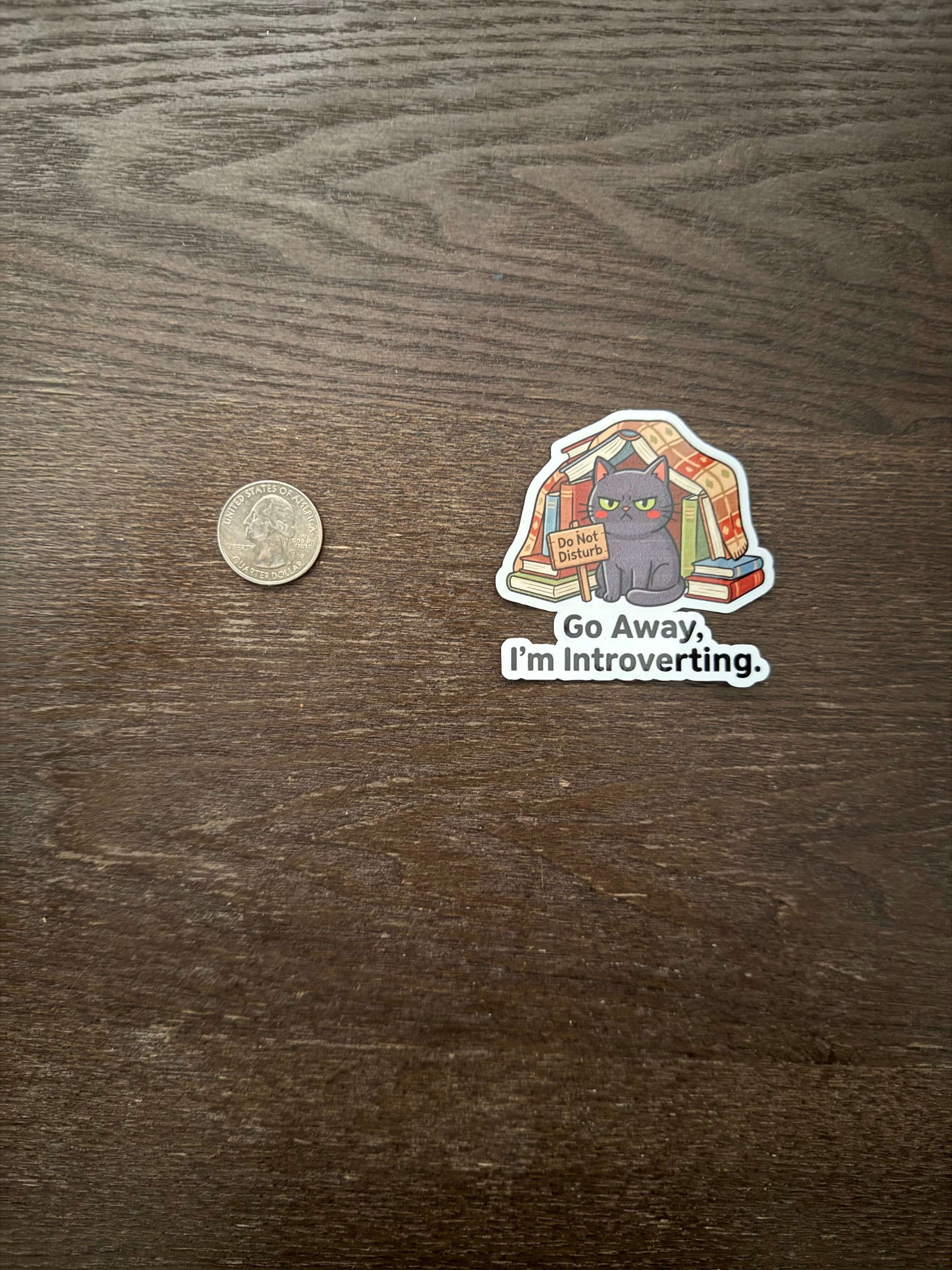Sticker with a cat illustration and text 'Go Away, I'm Introverting' on a wooden surface with a coin for scale.
