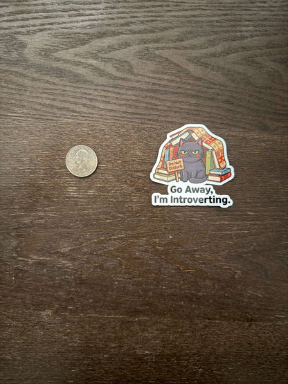Sticker with a cat illustration and text 'Go Away, I'm Introverting' on a wooden surface with a coin for scale.