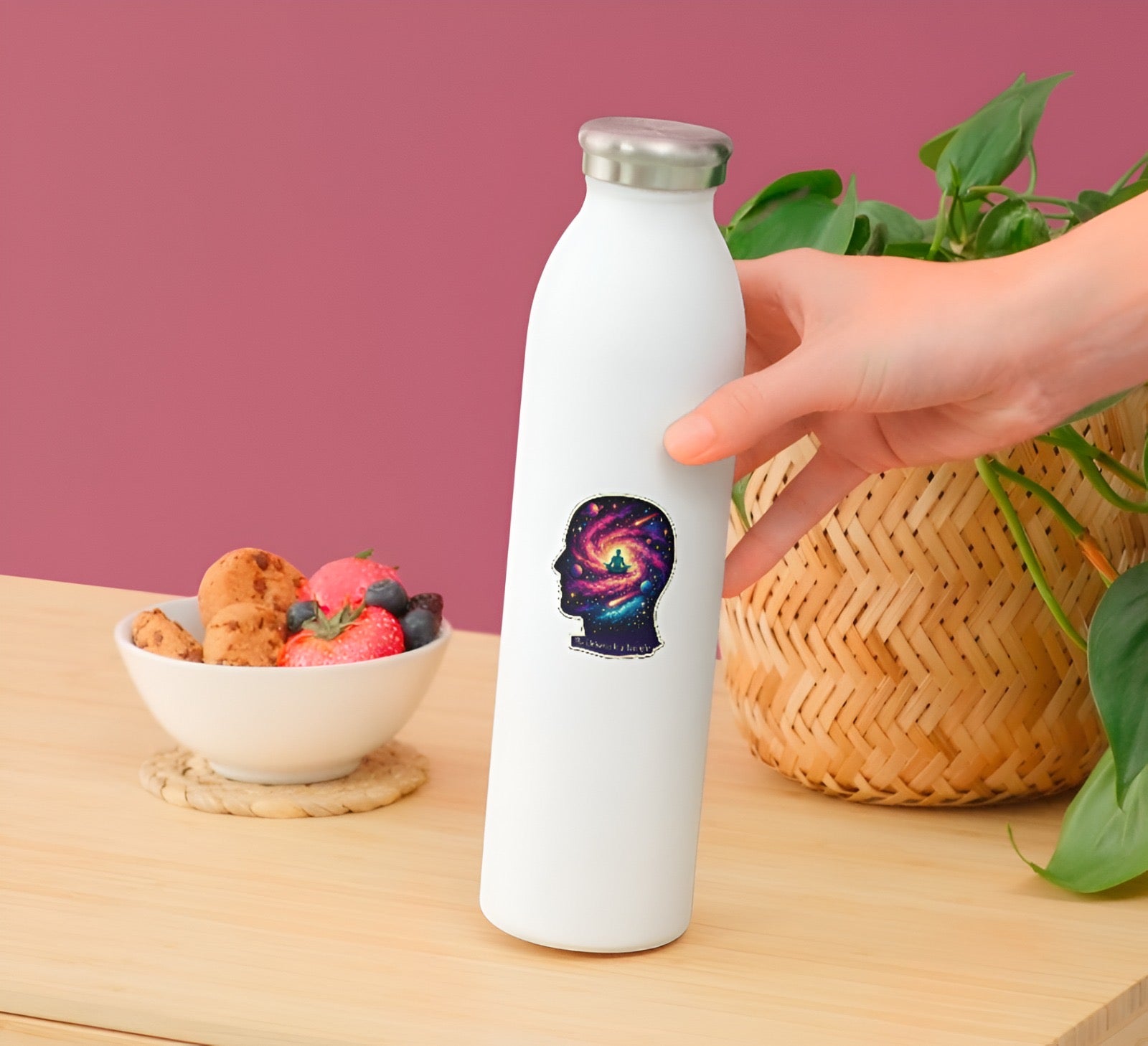 White water bottle with a colorful design held by a hand on a wooden table with a bowl of fruit and a plant in the background.