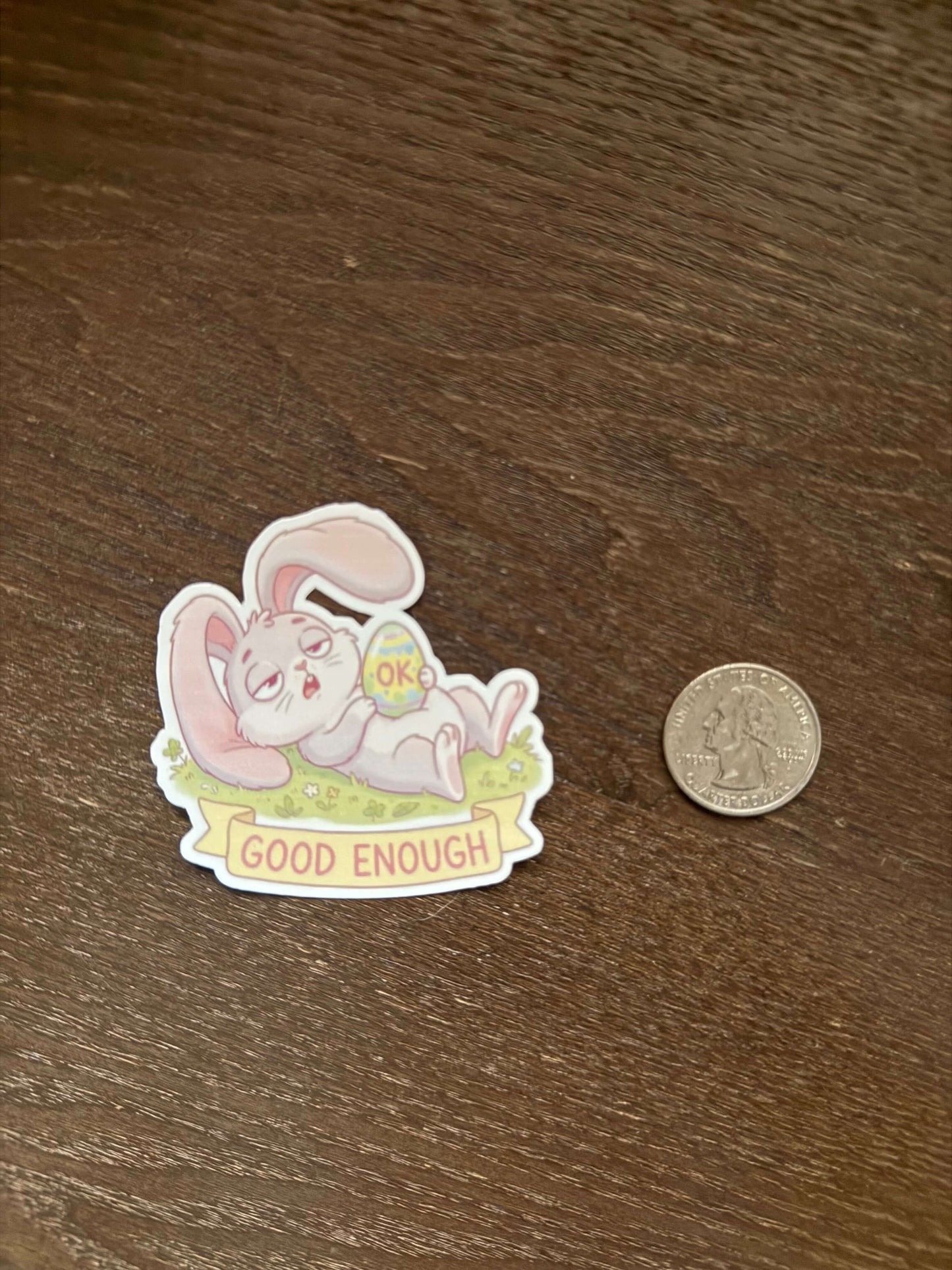 Sticker of a cartoon character with text on a wooden surface next to a coin for scale.