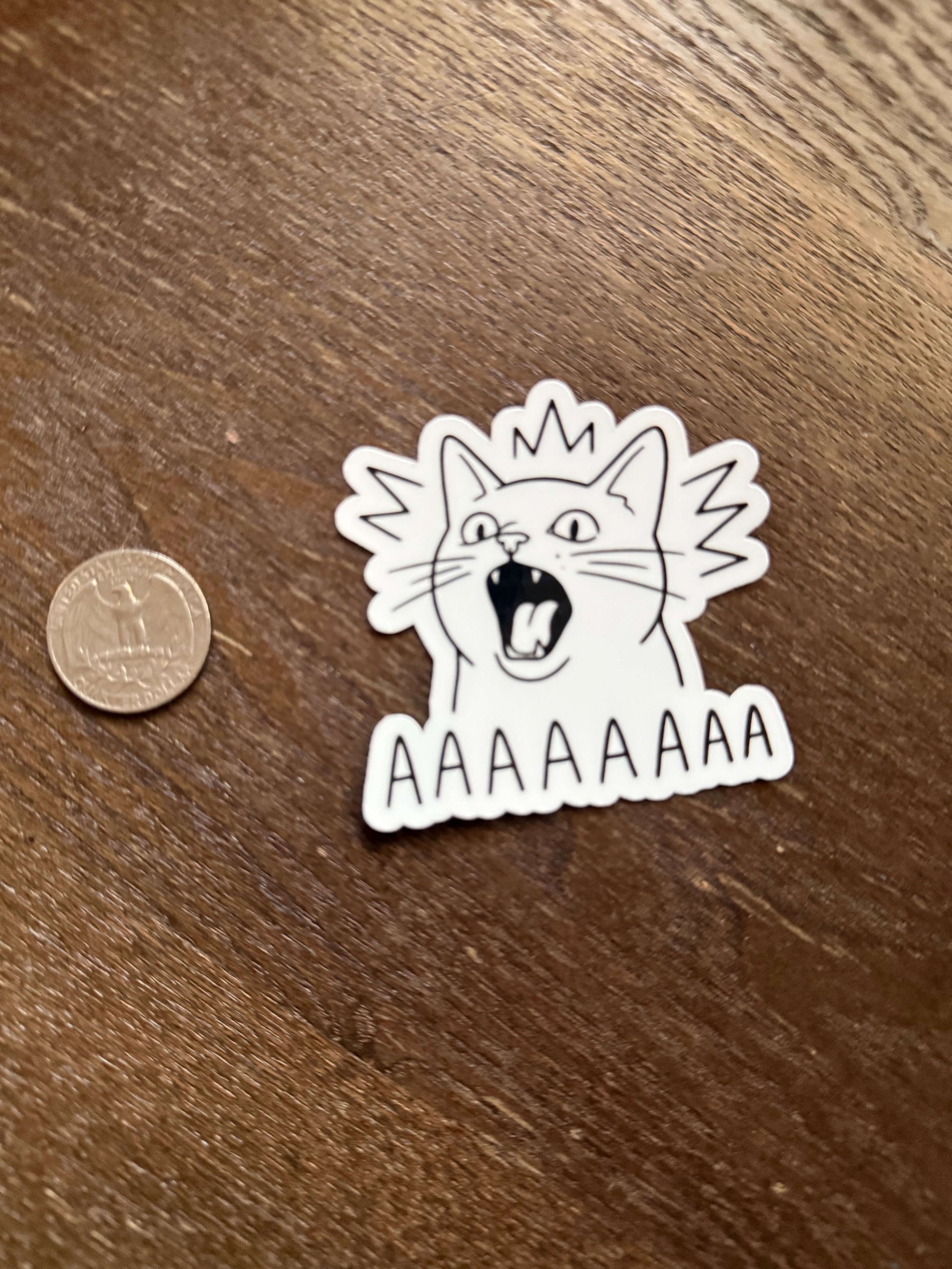 Sticker of a cat with a distressed expression on a wooden surface next to a coin for scale.
