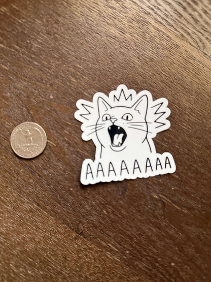 Sticker of a cat with a distressed expression on a wooden surface next to a coin for scale.