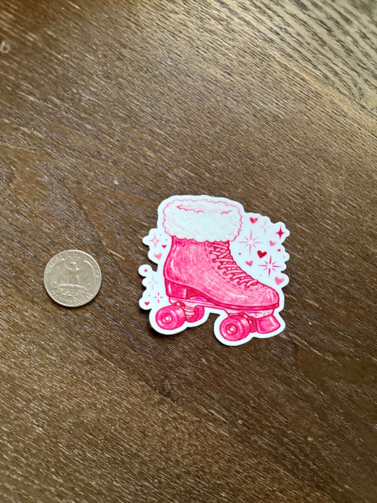 Pink roller skate sticker with white details on a wooden surface next to a coin for scale.