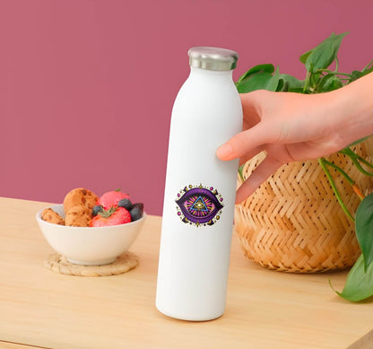 White water bottle with a colorful design held by a hand on a wooden table with a bowl of snacks and plant in the background.