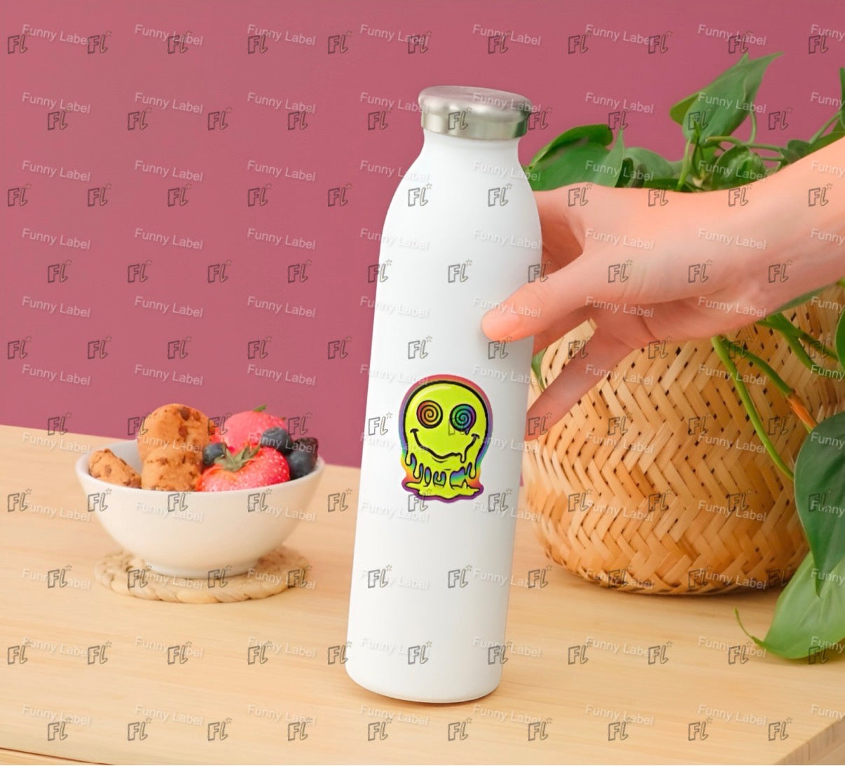 White water bottle with a smiley face design held by a hand on a wooden surface with a bowl of fruit and a basket in the background.