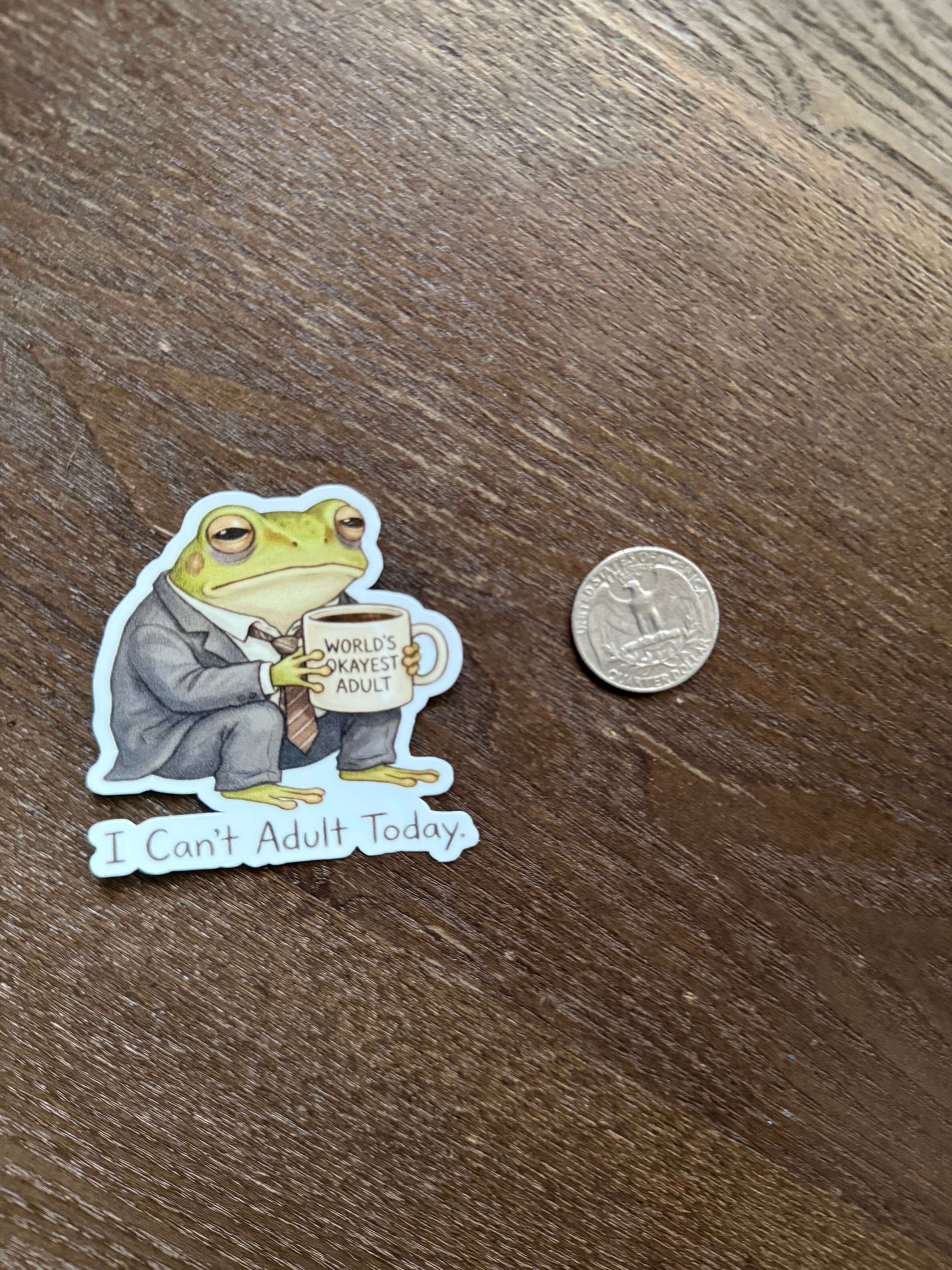 Sticker of a frog reading a book titled 'Don't Adult Today' next to a coin on a wooden surface