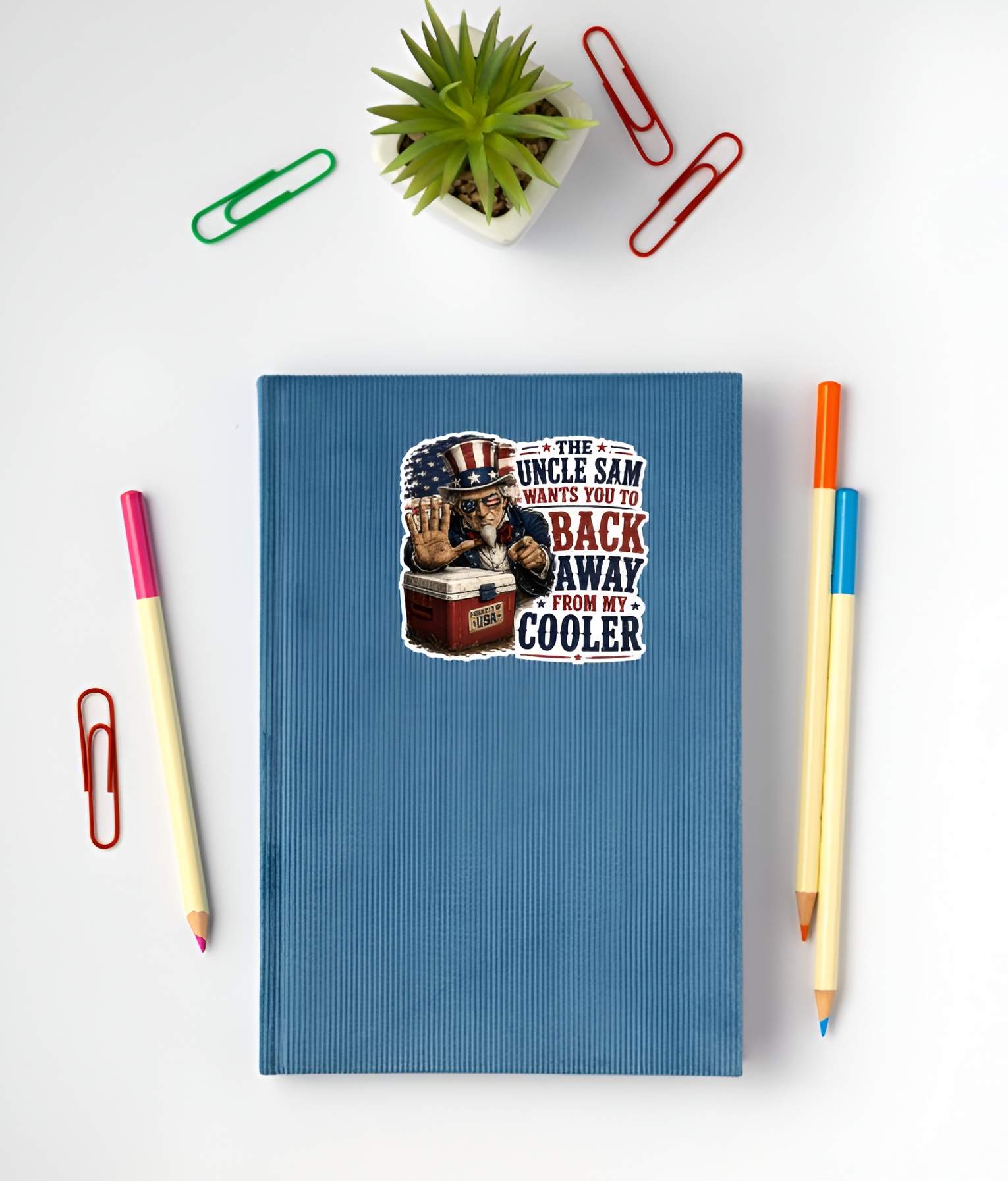 Blue notebook with a sticker featuring a detailed vintage woodcut illustration of an intense Uncle Sam wearing American flag sunglasses, holding up a stop hand over a red ice chest labeled "PROPERTY OF USA", next to bold red and blue text reading "THE UNCLE SAM WANTS YOU TO BACK AWAY FROM MY COOLER". on a white surface with stationery items.