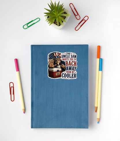 Blue notebook with a sticker featuring a detailed vintage woodcut illustration of an intense Uncle Sam wearing American flag sunglasses, holding up a stop hand over a red ice chest labeled "PROPERTY OF USA", next to bold red and blue text reading "THE UNCLE SAM WANTS YOU TO BACK AWAY FROM MY COOLER". on a white surface with stationery items.