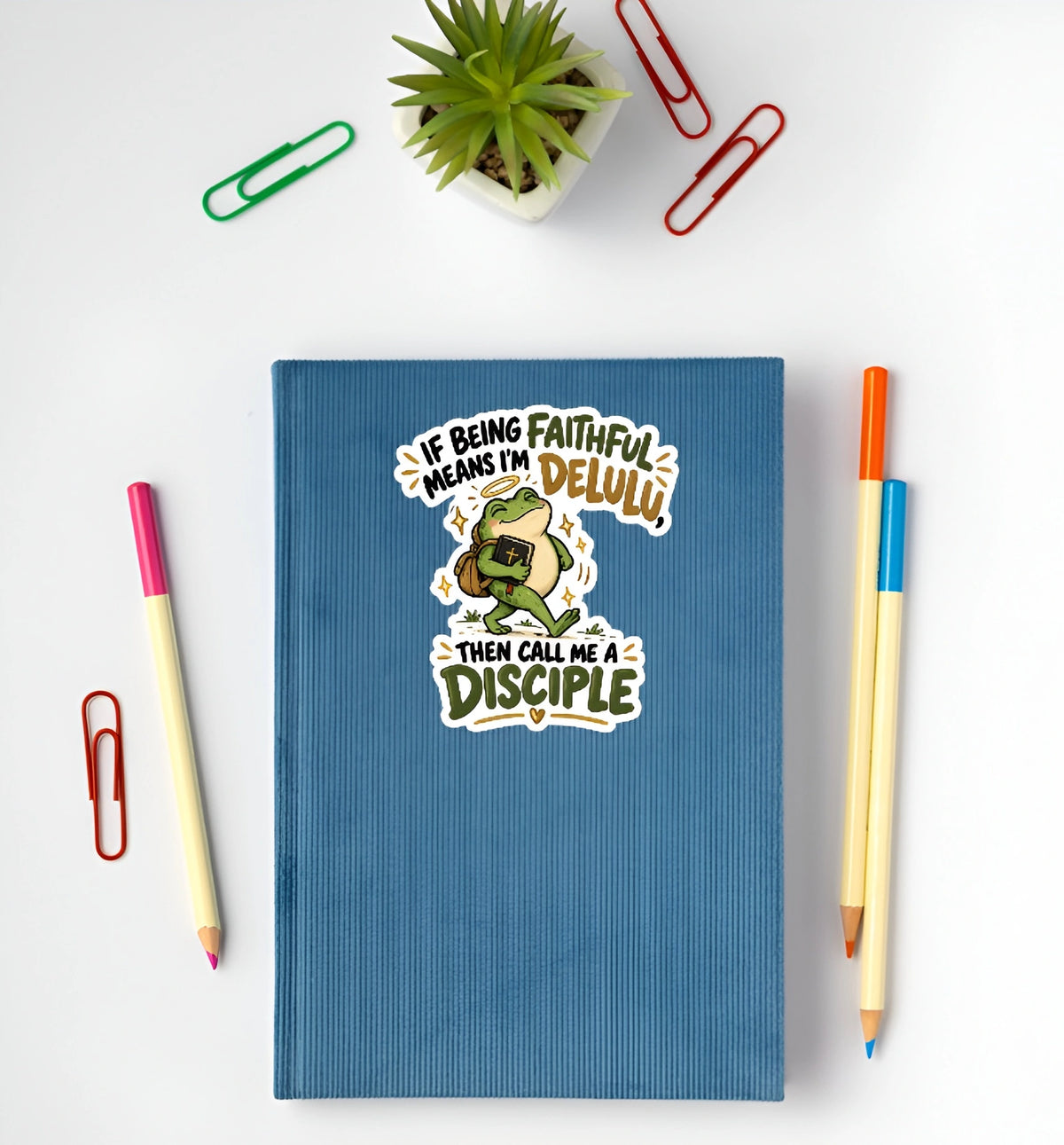 A blue textured notebook with a cartoon frog and the text 'IF BEING FAITHFUL MEANS I'M DELULI THEN CALL ME A DISCIPLE' lies on a white surface, surrounded by colored pencils and paperclips.