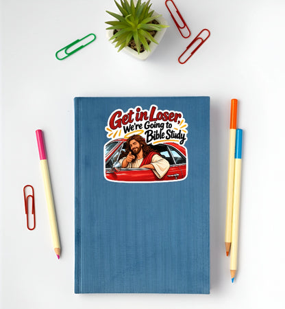 A blue hardcover notebook with a cartoon graphic of Jesus in a red car and the text 'Get in Loser We're Going to Bible Study', styled in a flat-lay with paper clips, pencils, and a small plant on a white background.