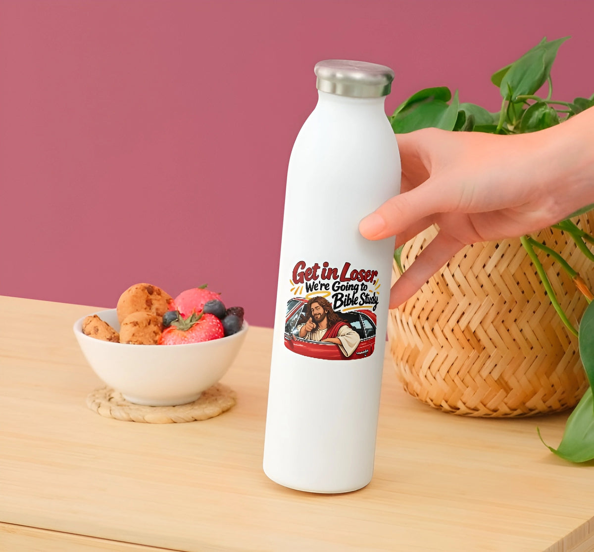 A hand holds a white insulated water bottle with a silver cap on a wooden table. The bottle has a colorful graphic that reads 'Get in Loser We're Going to Bible Study' with an illustration.
