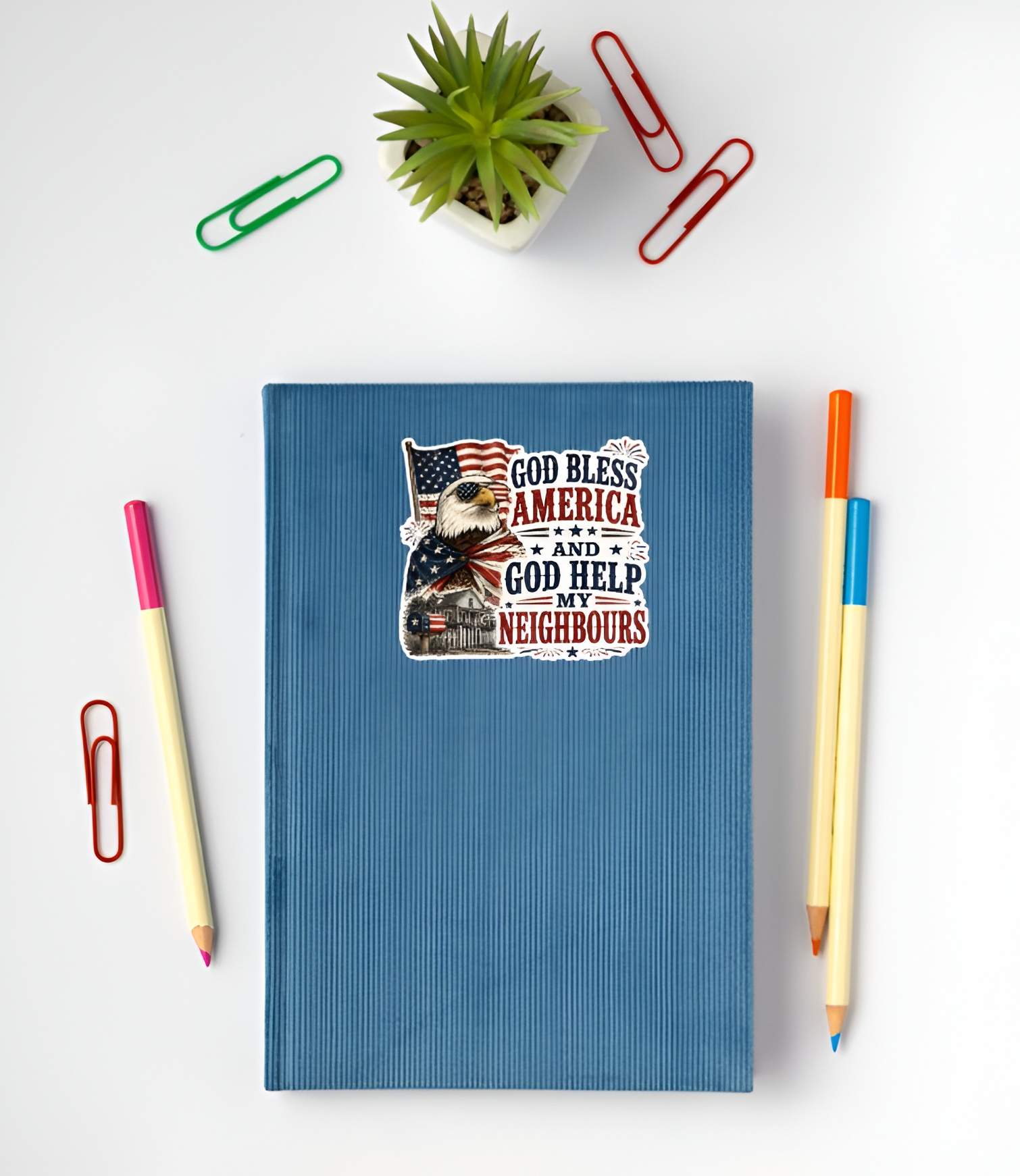 Blue notebook with a printed design featuring a patriotic detailed illustration of a bald eagle wearing American flag sunglasses and draped in a flag cape, standing next to a house with a flag mailbox under a waving US flag, paired with bold text reading "GOD BLESS AMERICA AND GOD HELP MY NEIGHBOURS" on a white surface with stationery items.