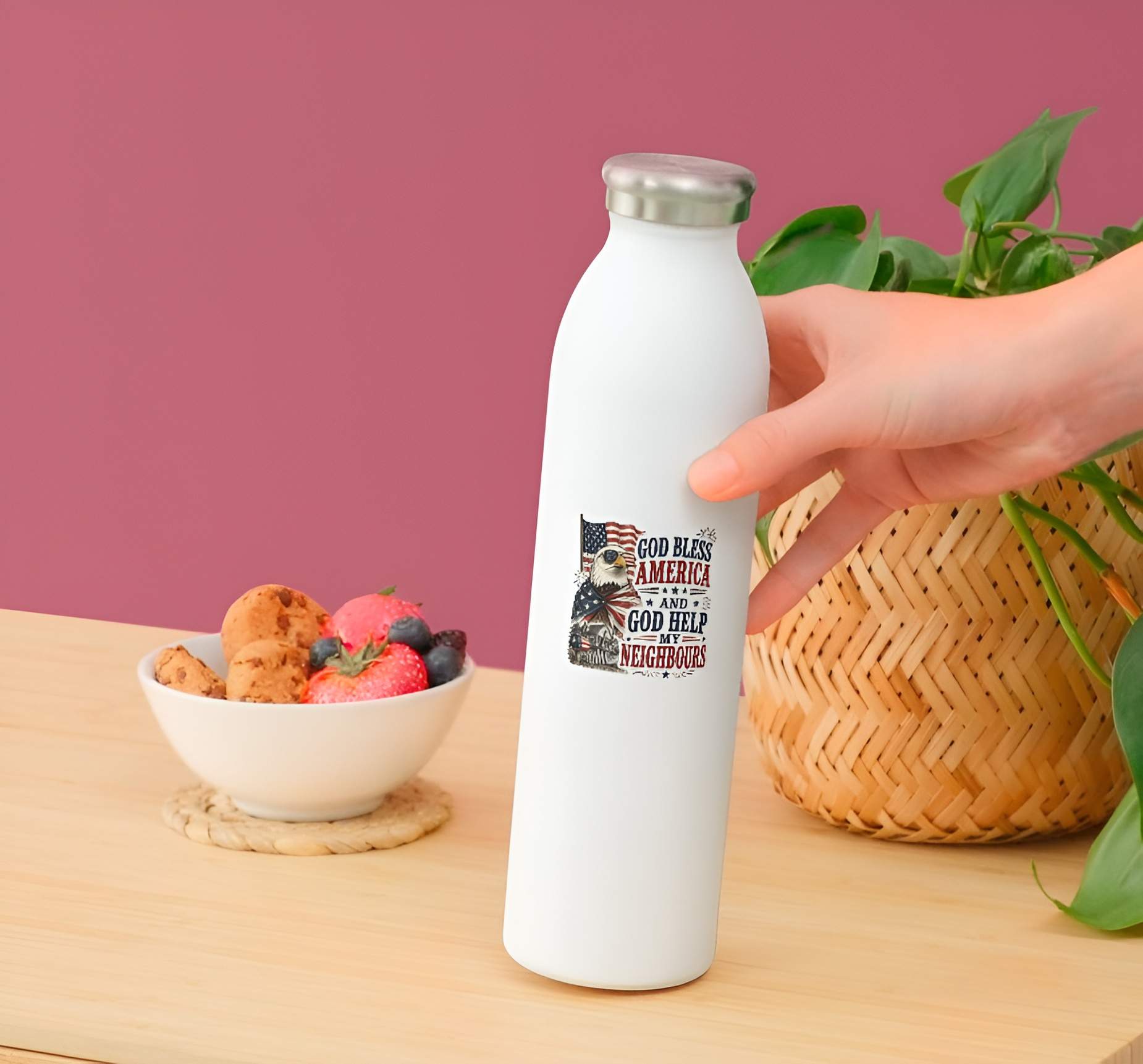 White water bottle with a printed design featuring a patriotic detailed illustration of a bald eagle wearing American flag sunglasses and draped in a flag cape, standing next to a house with a flag mailbox under a waving US flag, paired with bold text reading "GOD BLESS AMERICA AND GOD HELP MY NEIGHBOURS".held by a hand on a wooden table with a bowl of snacks and a plant.
