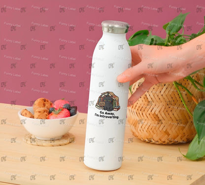 White water bottle with a design and text held by a hand on a table with a bowl of fruit and a plant.
