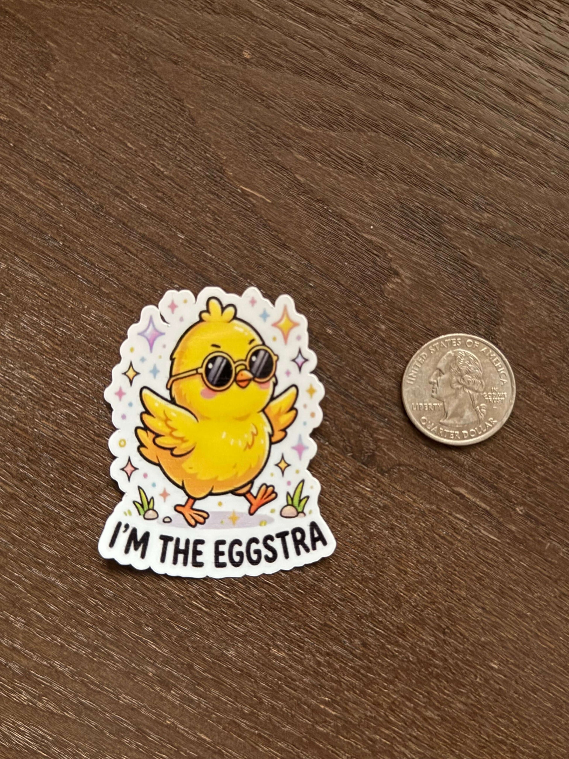 Sticker of a yellow chick with sunglasses labeled 'I'm the Eggstra' next to a coin on a wooden surface