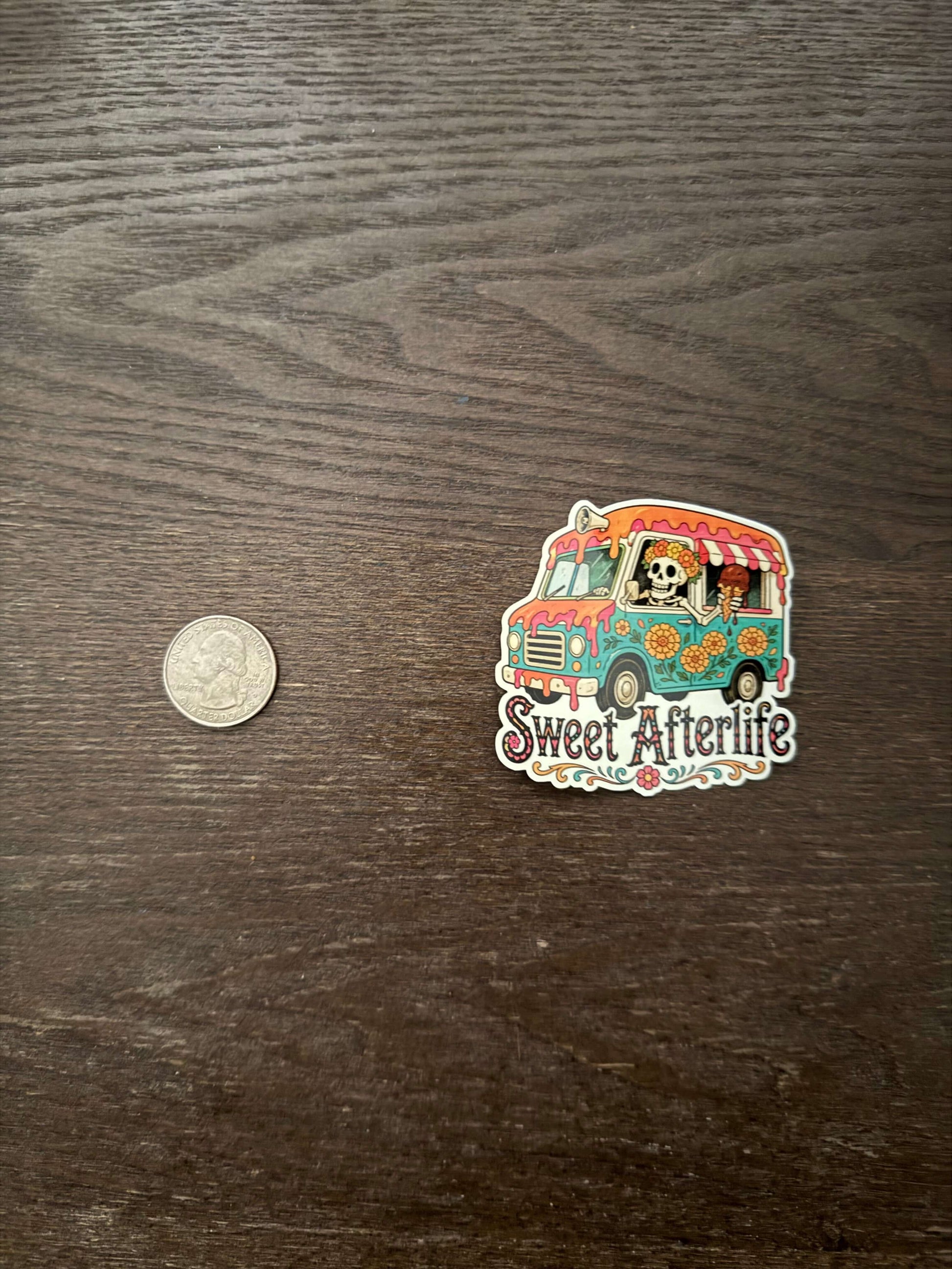 Colorful 'Sweet Afterlife' sticker on a wooden surface with a coin for scale.