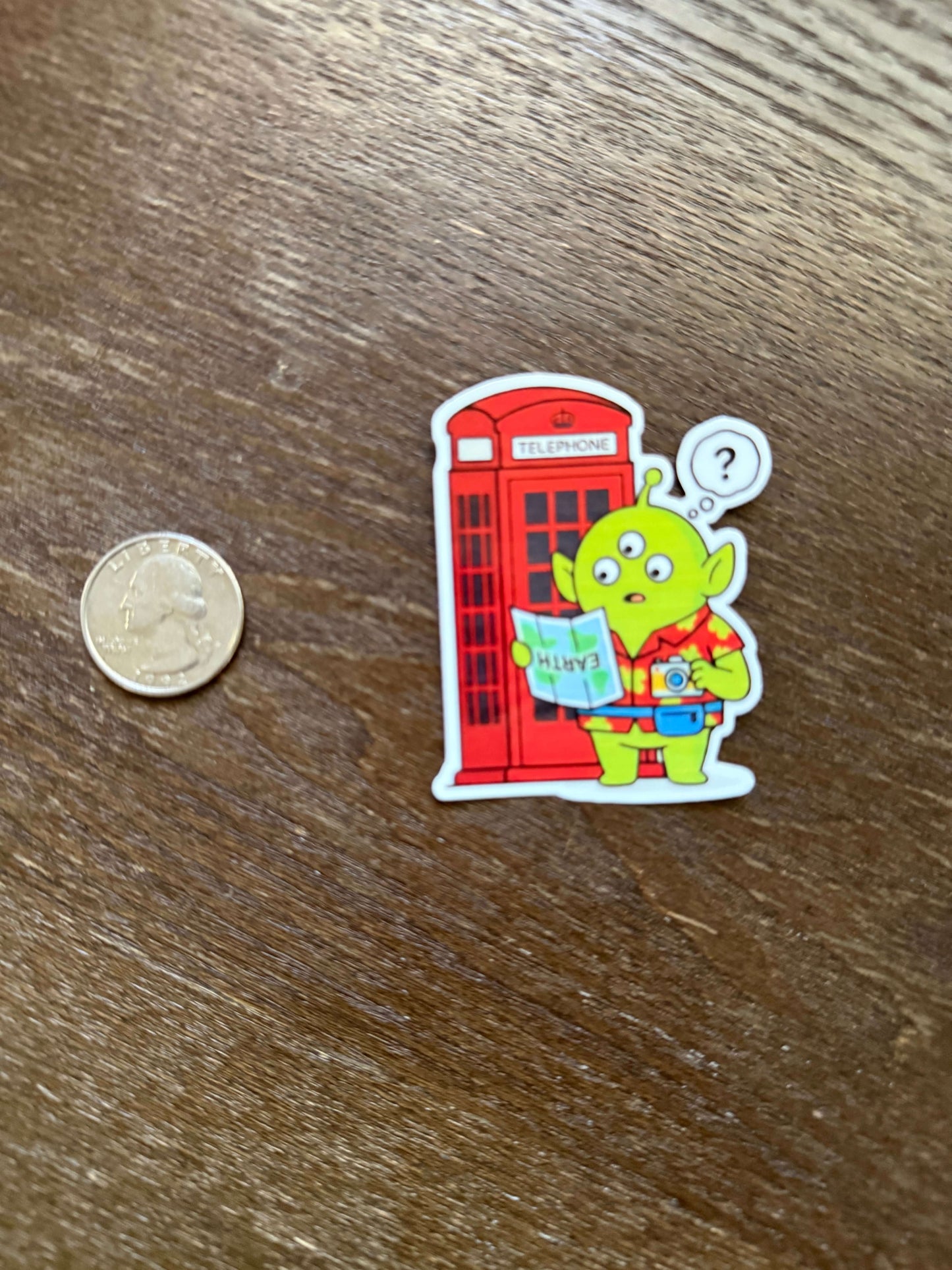 Sticker of a green alien in a red telephone box on a wooden surface with a coin for scale.
