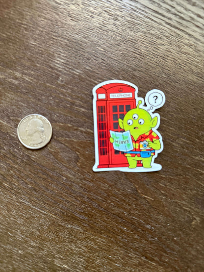 Sticker of a green alien in a red telephone box on a wooden surface with a coin for scale.