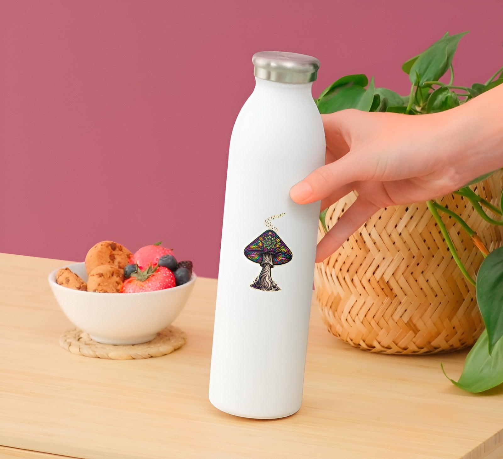 White water bottle with a mushroom design held by a hand on a wooden table with a bowl of fruit and a plant in the background.