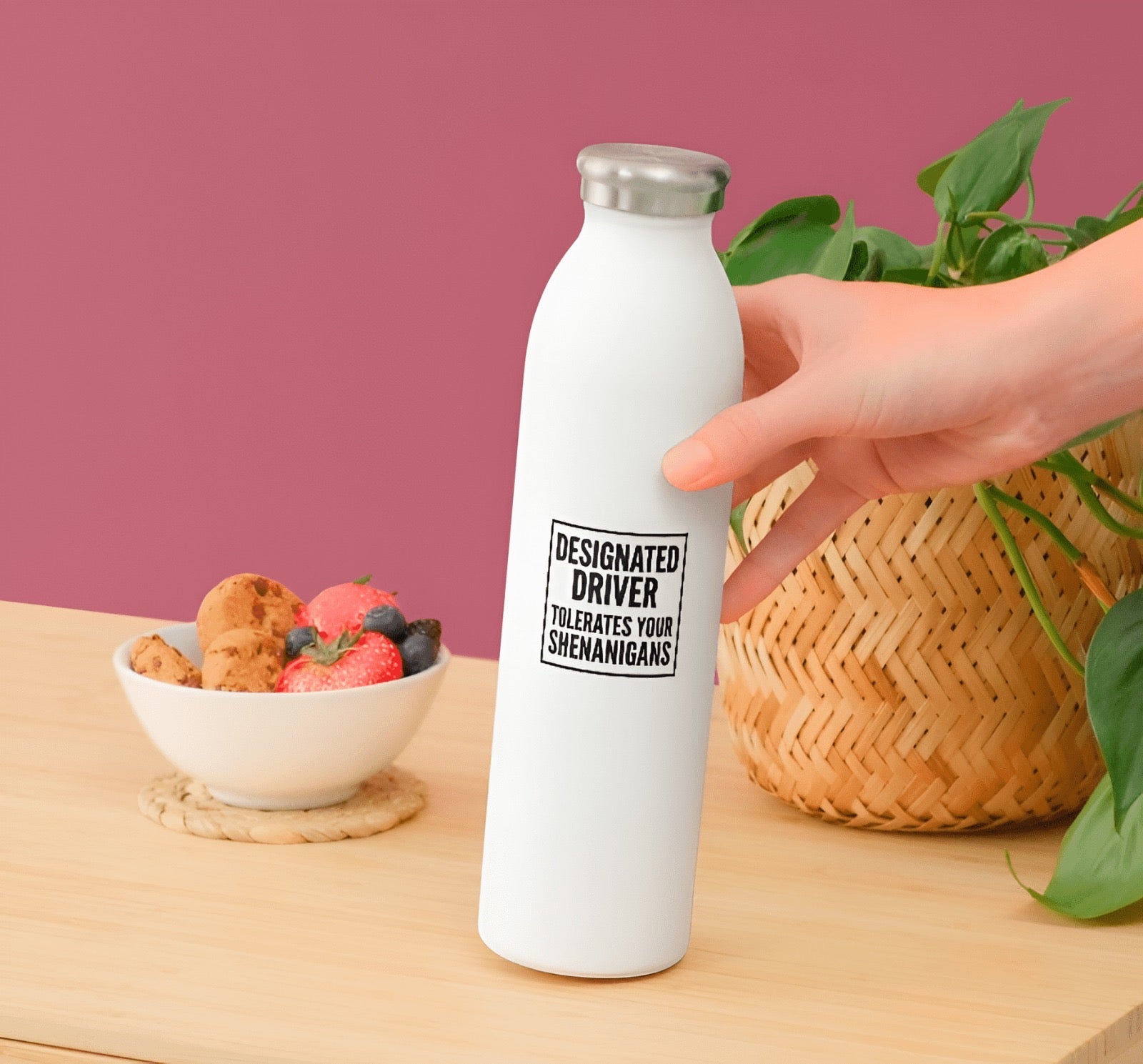 White water bottle with a humorous message held by a hand on a wooden surface with a bowl of snacks and plant in the background.