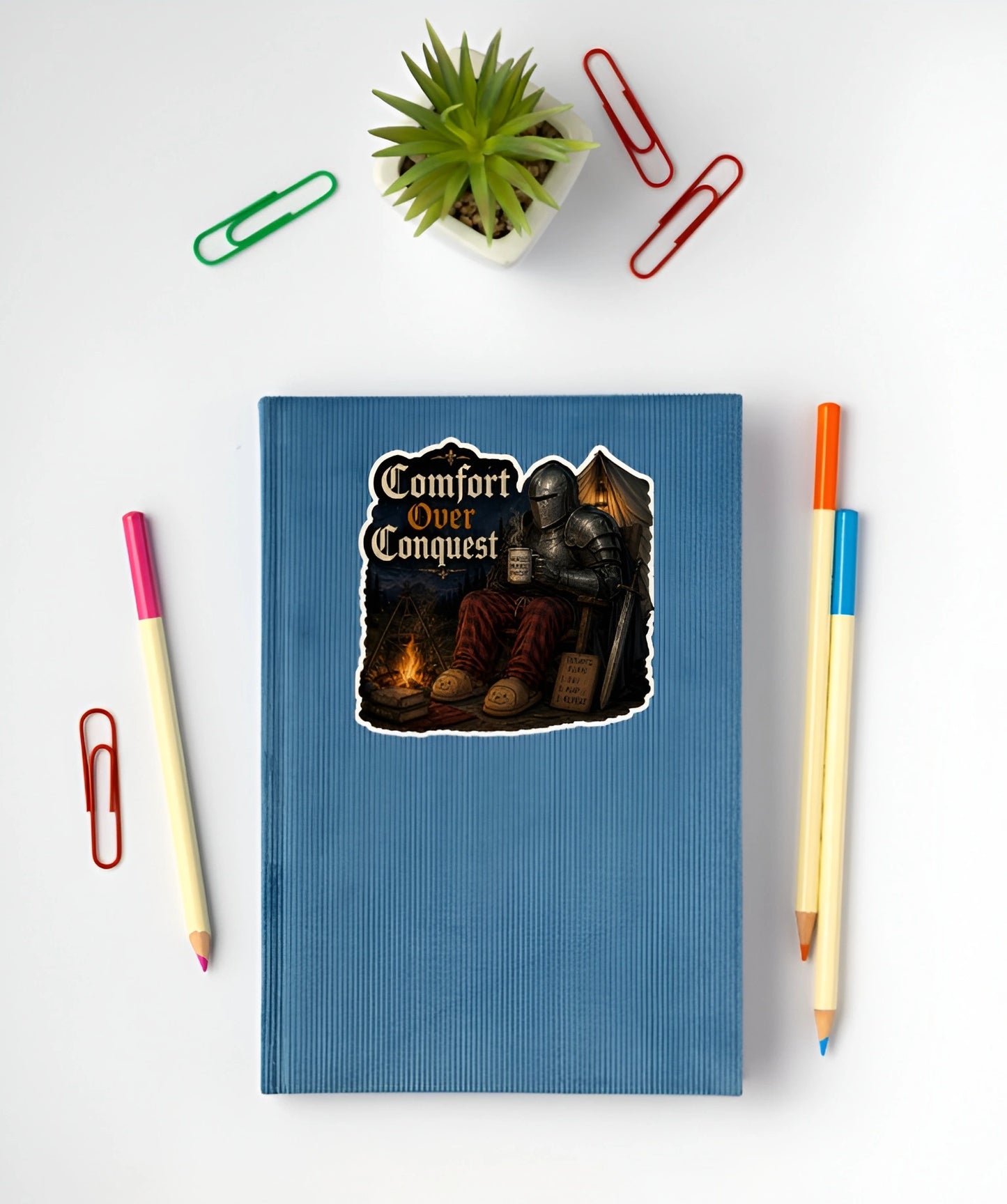 Comfort Over Conquest medieval knight sticker with plaid pants and campfire displayed on a blue notebook