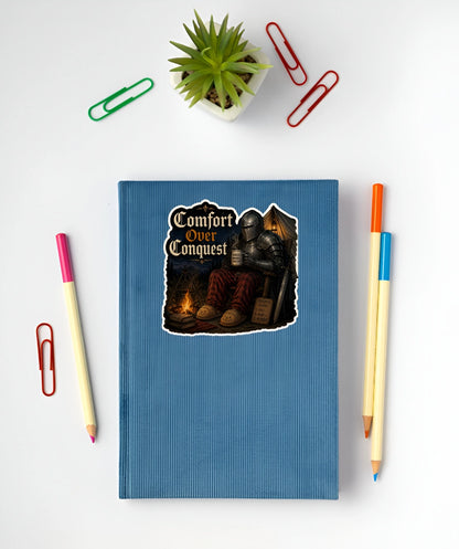 Comfort Over Conquest medieval knight sticker with plaid pants and campfire displayed on a blue notebook