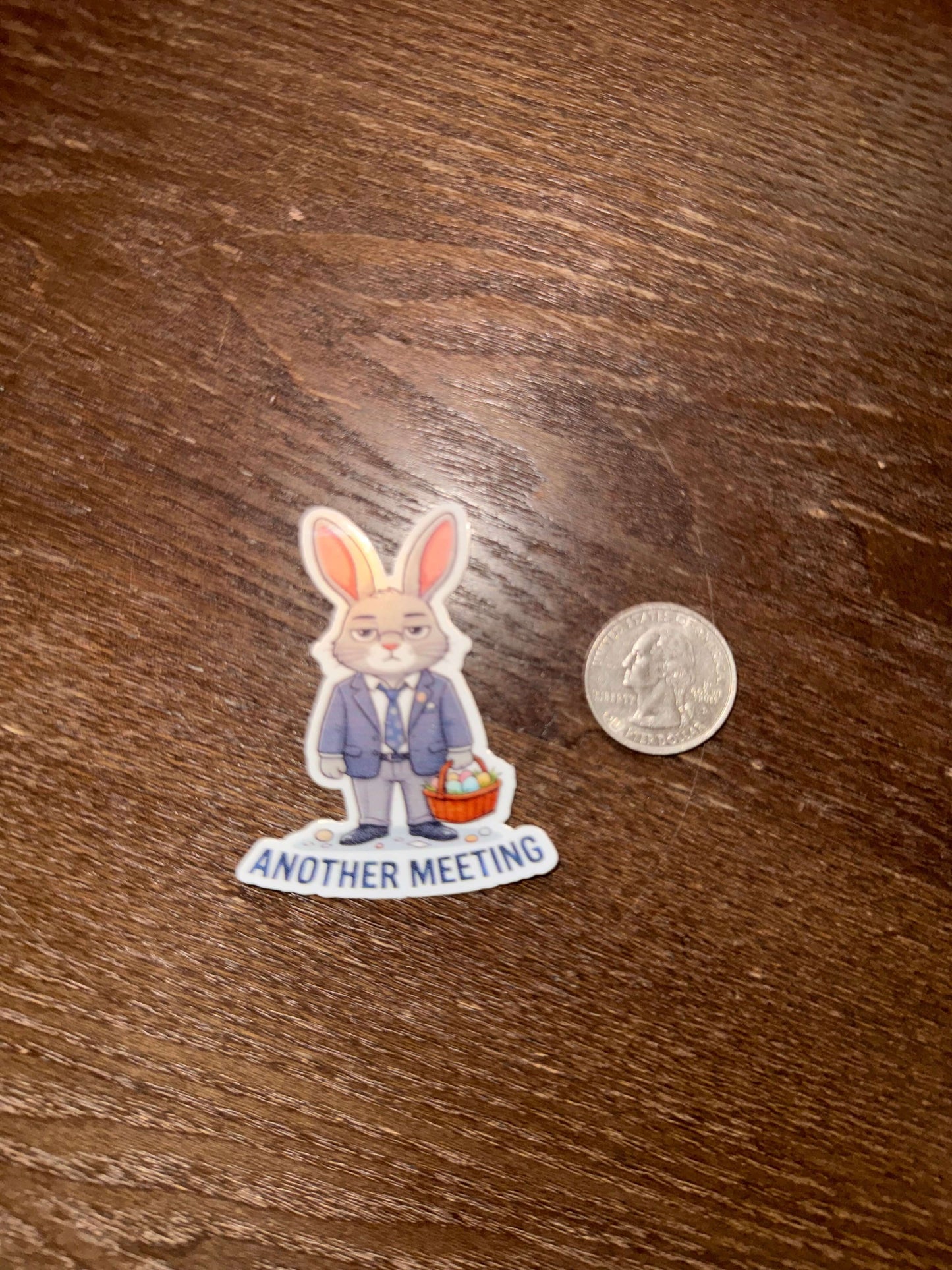 Sticker of a rabbit and person with text 'ANOTHER MEETING' next to a quarter on a wooden surface