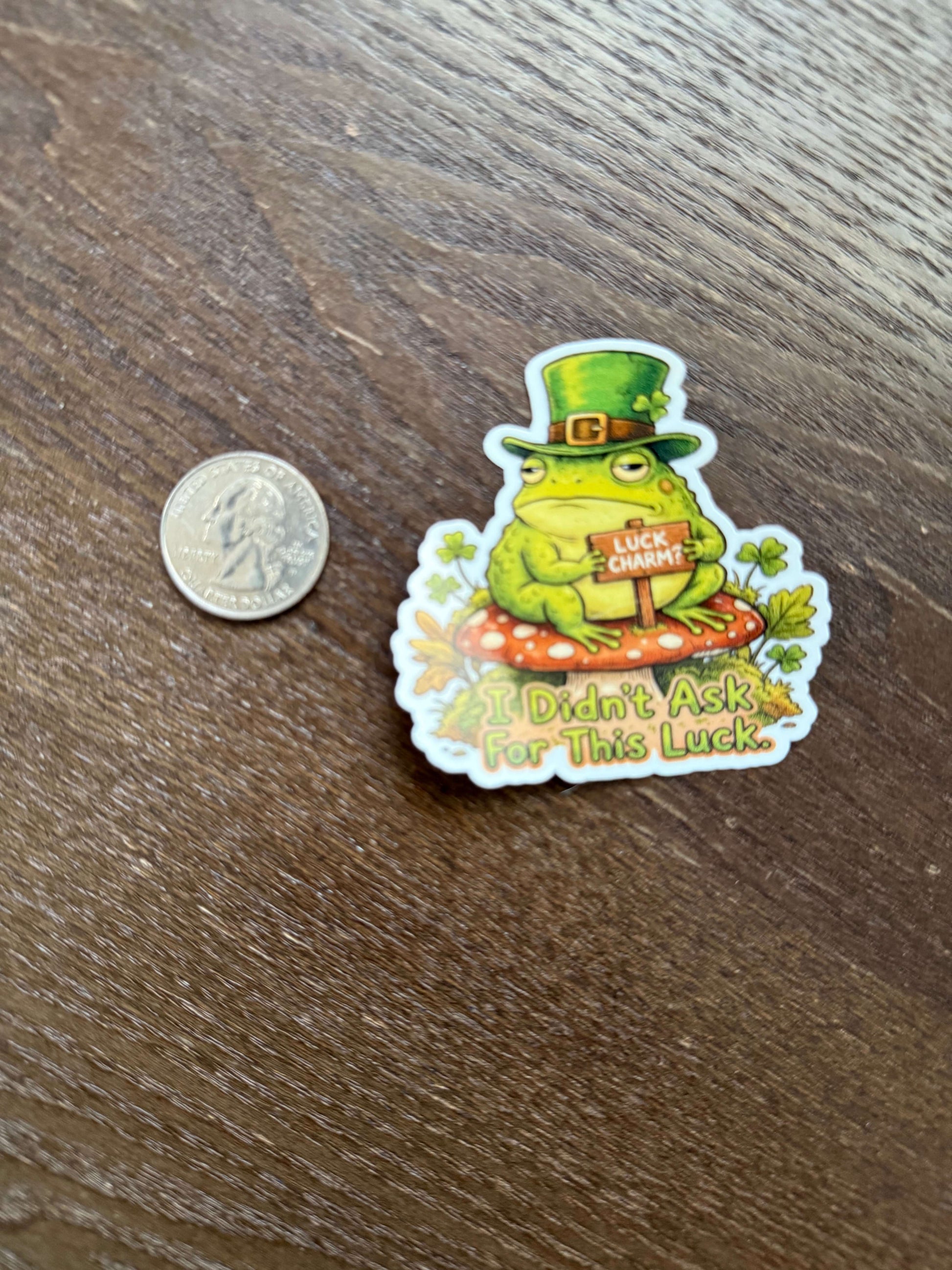 Sticker of a frog wearing a top hat with text on a wooden surface next to a coin for size reference.