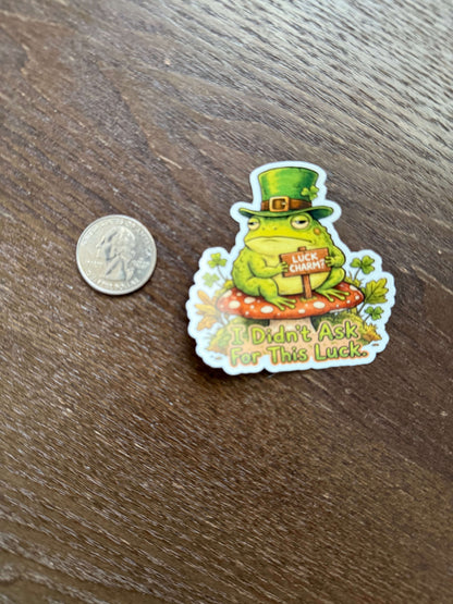 Sticker of a frog wearing a top hat with text on a wooden surface next to a coin for size reference.
