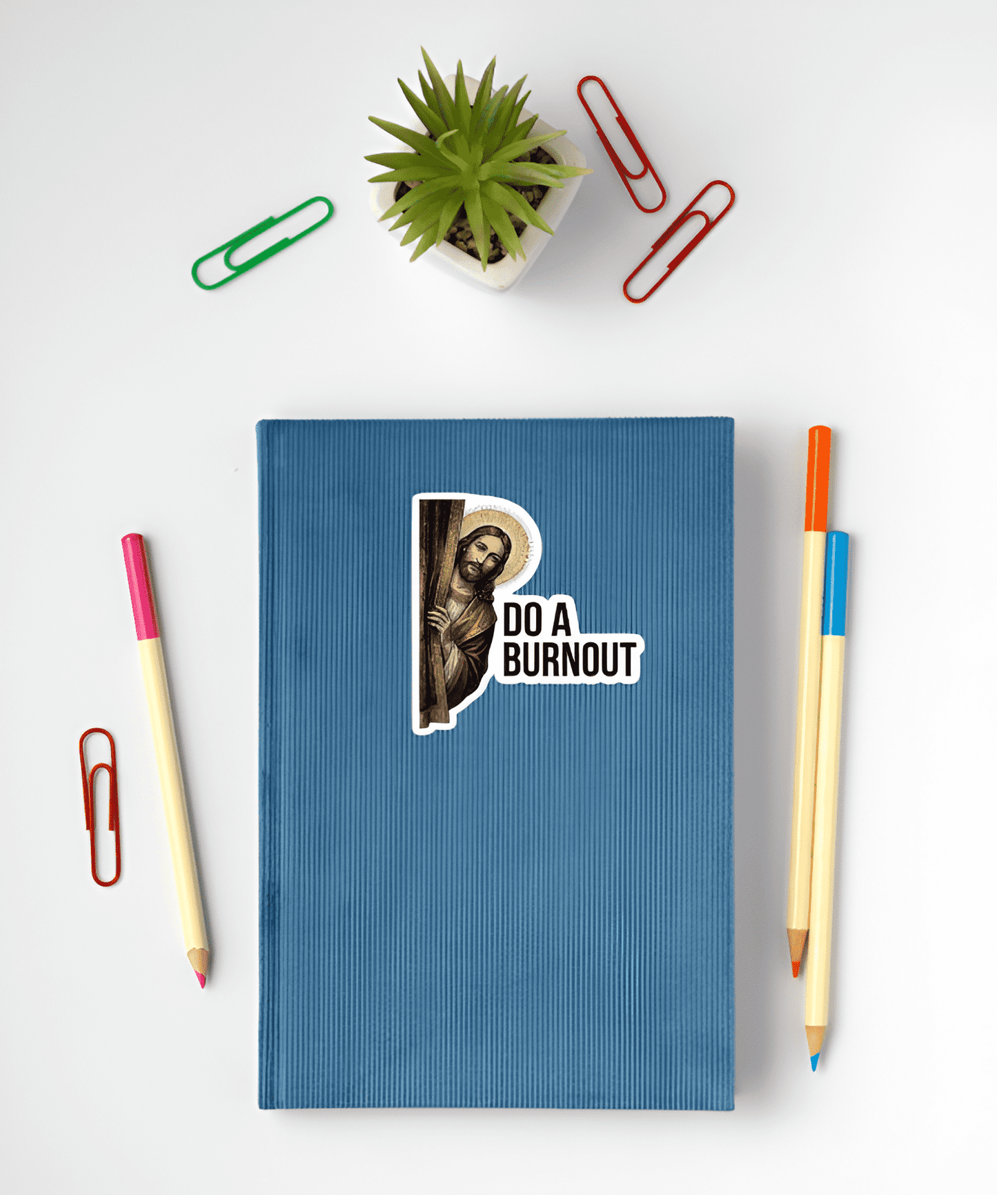 Blue notebook with  illustration of Jesus with a detailed golden halo, peeking out from behind a dark burgundy curtain and looking directly outward next to bold black text reading "DO A BURNOUT". sticker on a white surface with stationery items.