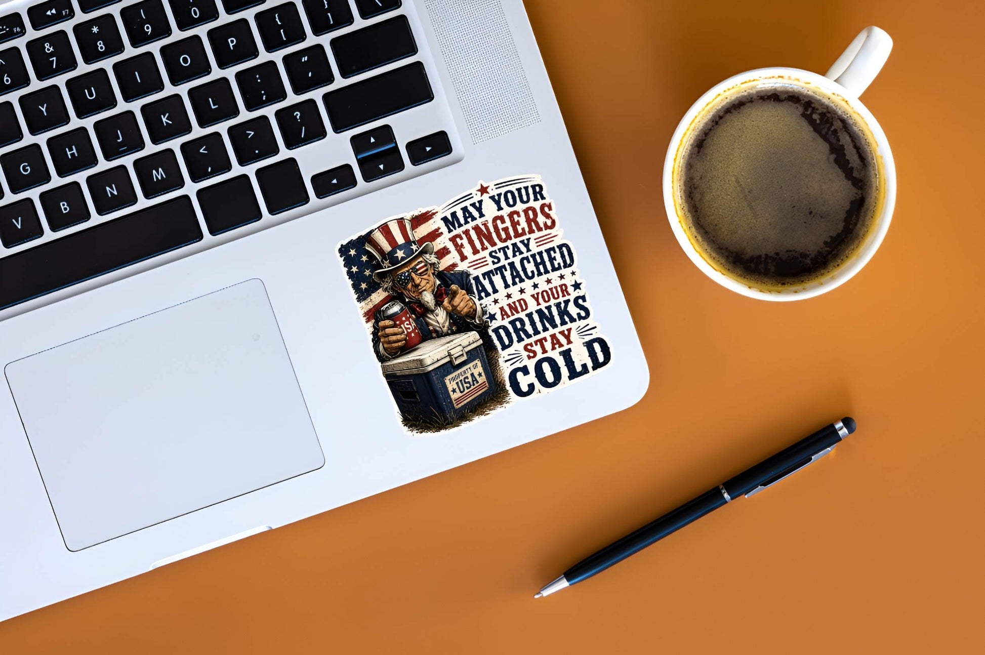 Laptop with a humorous sticker featuring a detailed vintage woodcut illustration of Uncle Sam wearing flag sunglasses, holding a USA beverage can next to a blue ice chest labeled "PROPERTY OF USA", alongside bold red and blue text reading "MAY YOUR FINGERS STAY ATTACHED AND YOUR DRINKS STAY COLD". on a desk next to a cup of coffee and pen