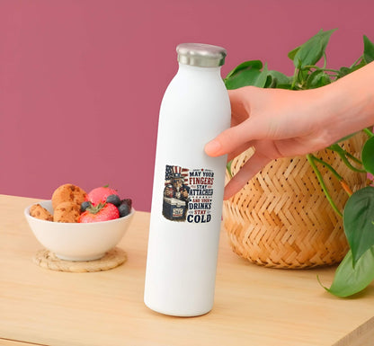 White water bottle with a sticker featuring a detailed vintage woodcut illustration of Uncle Sam wearing flag sunglasses, holding a USA beverage can next to a blue ice chest labeled "PROPERTY OF USA", alongside bold red and blue text reading "MAY YOUR FINGERS STAY ATTACHED AND YOUR DRINKS STAY COLD".held by a hand on a wooden table with a bowl of fruit and a plant.