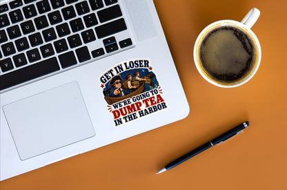 A die-cut sticker with a nautical illustration and the text 'GET IN LOSER WE'RE GOING TO DUMP TEA IN THE HARBOR' applied to the palm rest of a silver laptop. The laptop is shown in a flat-lay arrangement on a warm orange background, alongside a cup of black coffee and a black pen.